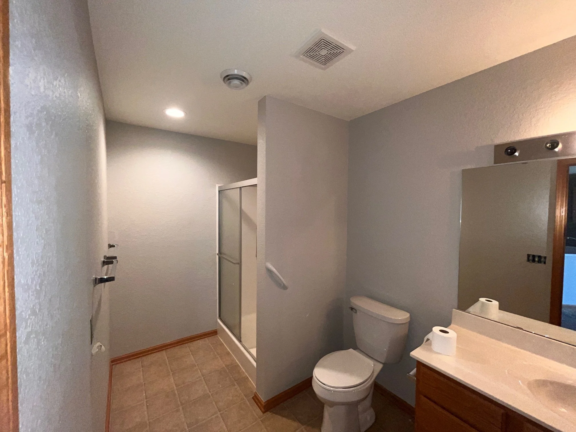 Bathroom with a toilet, vanity with a mirror, two rolls of toilet paper, a shower with frosted glass door, beige floor tiles, gray walls, ceiling vent, and recessed lighting.