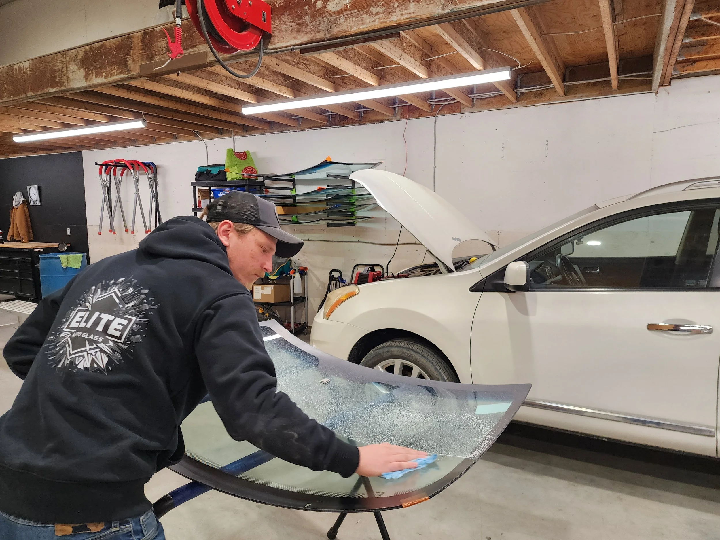 A man cleaning a large glass car windshield in a garage with a white car and an open hood in the background.