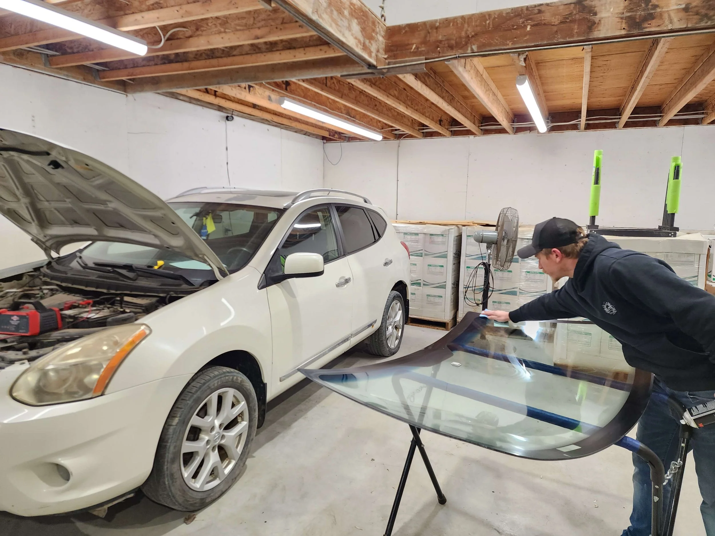 A man inspecting a car windshield in a garage with an open hood. The garage has wooden beams on the ceiling and fluorescent lighting.