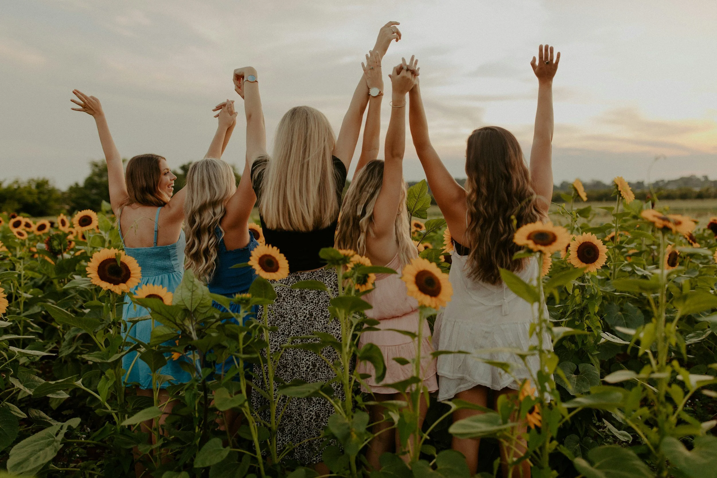 Six women in summer dresses are dancing with their hands raised in a sunflower field at sunset.
