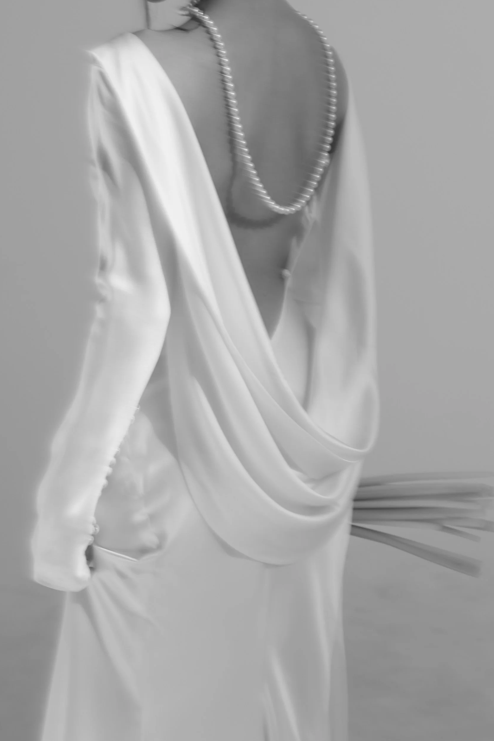 A person wearing elegant white satin clothing with a deep back neckline, accessorized with a pearl necklace.