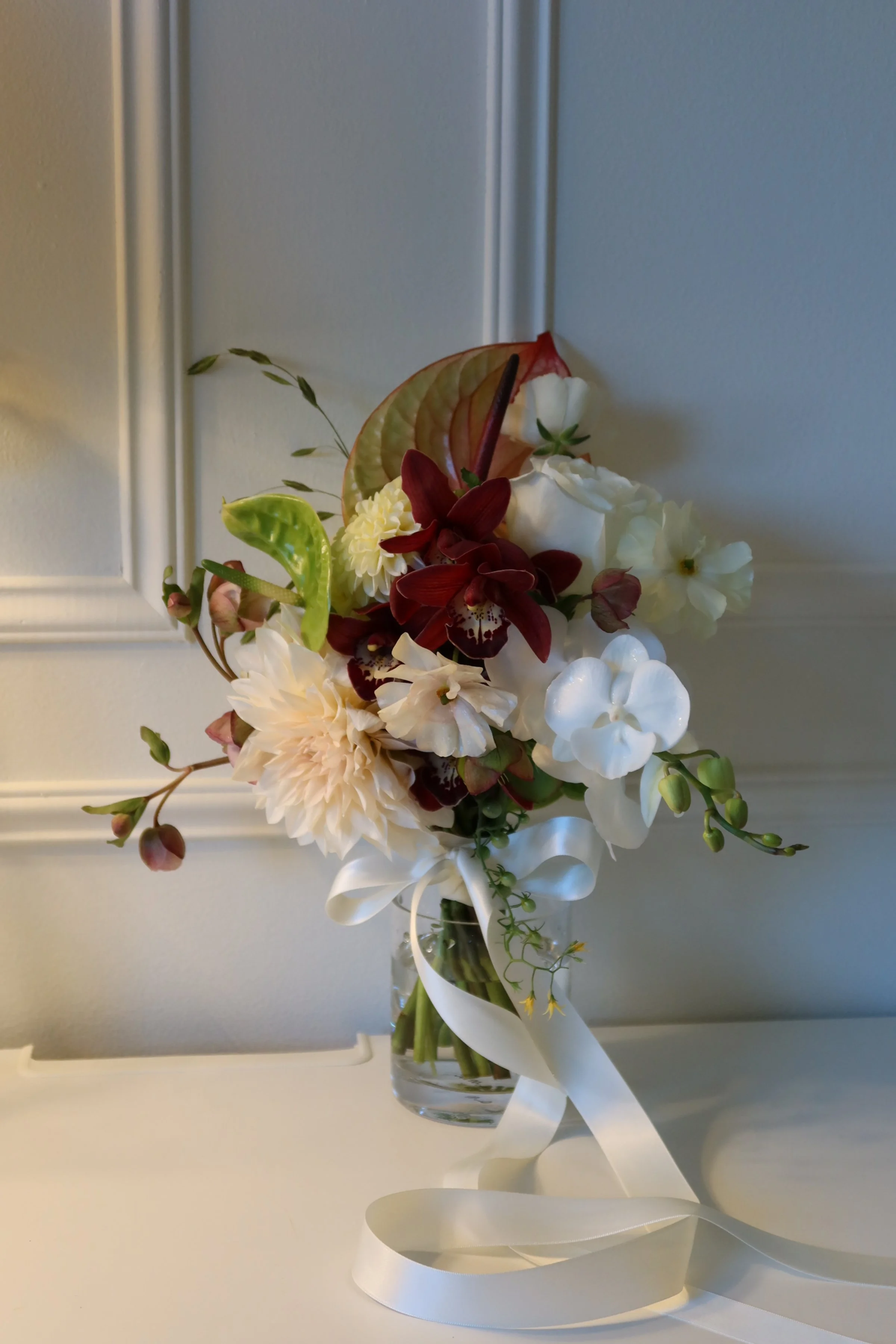A bouquet of flowers in a glass vase with white, cream, red, and green blooms and leaves, tied with a white ribbon.