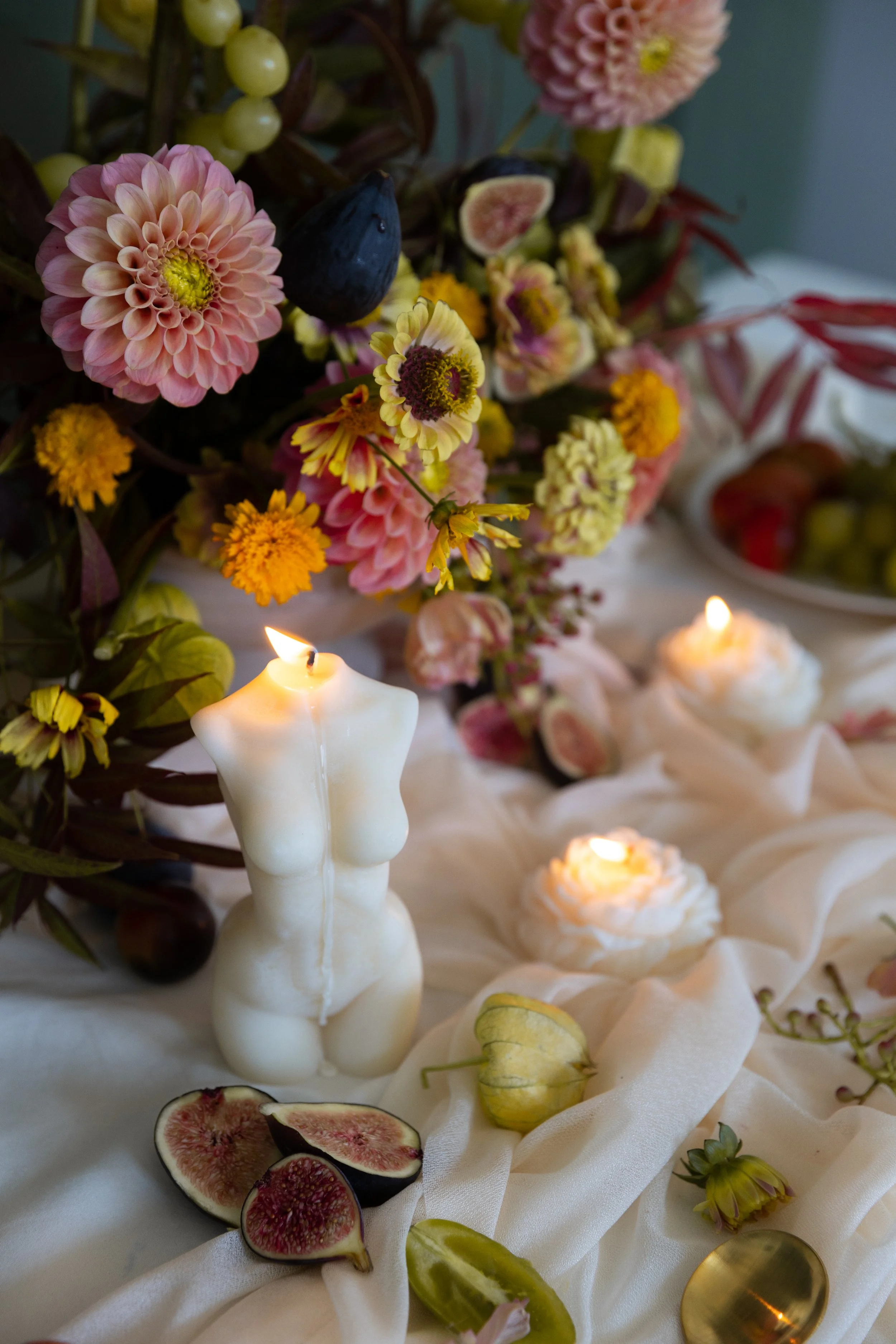 A table display featuring a woman-shaped candle, flowers, figs, and candles on white fabric with a bowl of fruit in the background.