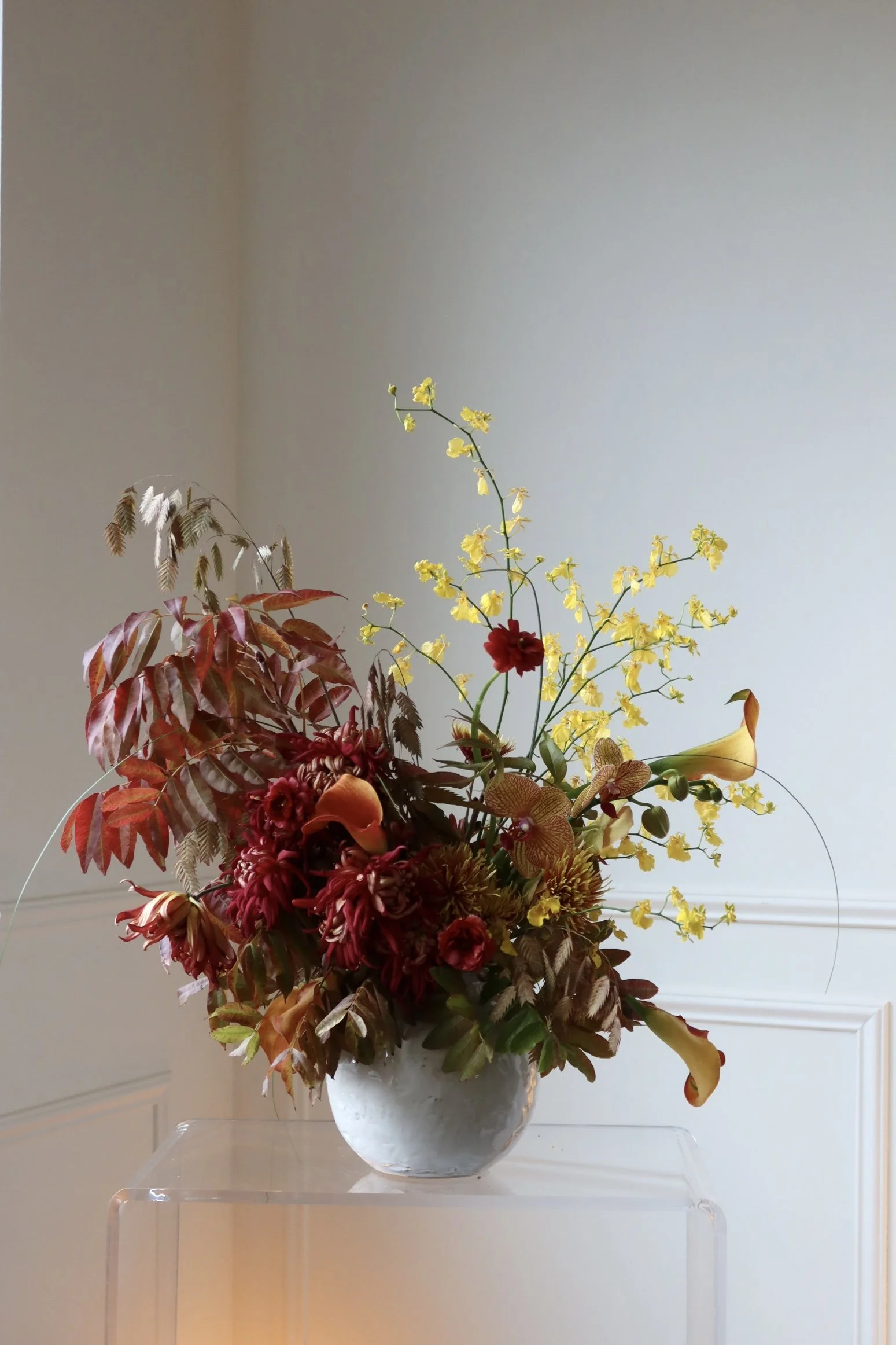 A floral arrangement with various colorful flowers and foliage in a white ceramic vase, displayed on a transparent stand against a plain wall.