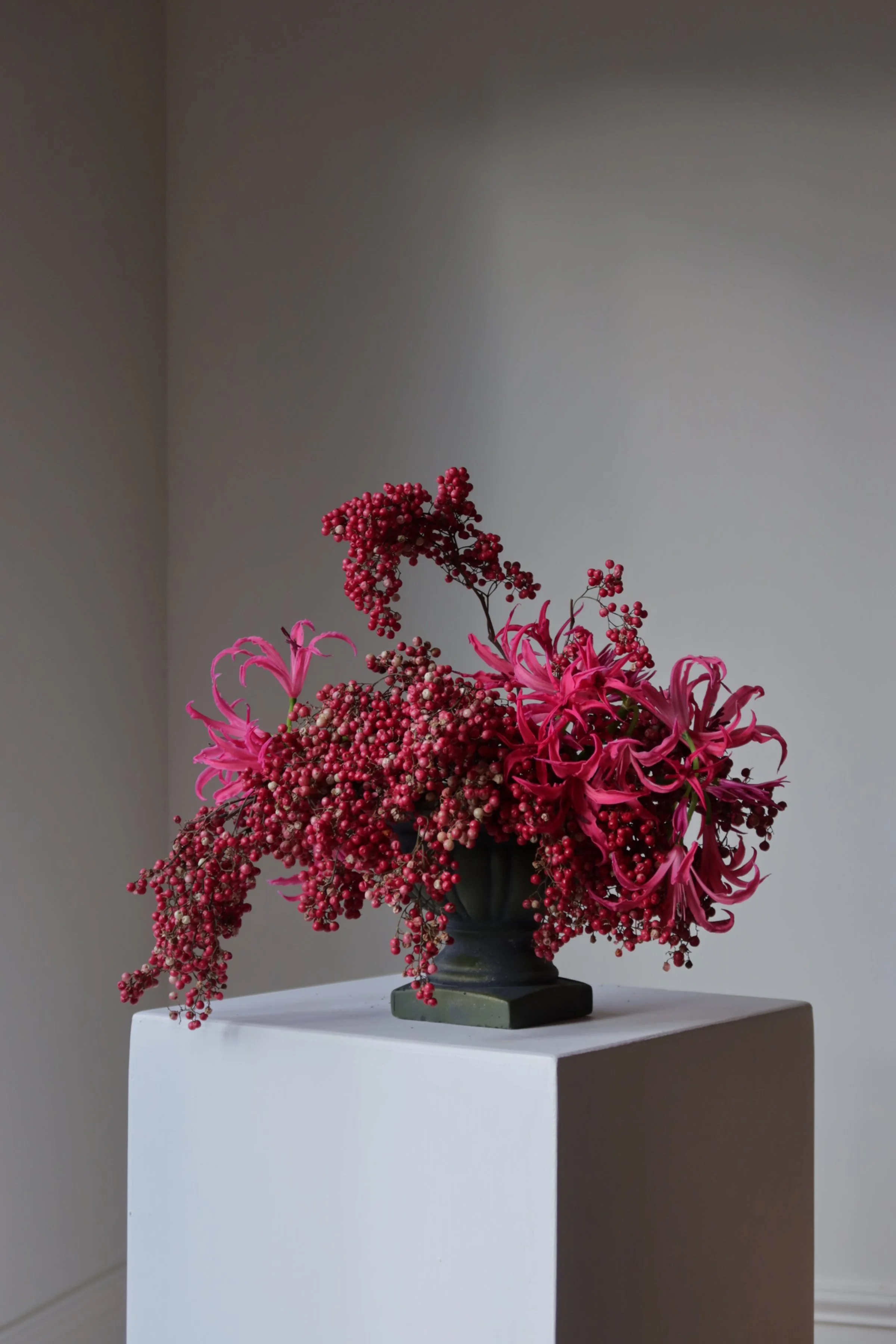 Pink and red floral arrangement in a dark vase on a white pedestal against a plain wall.