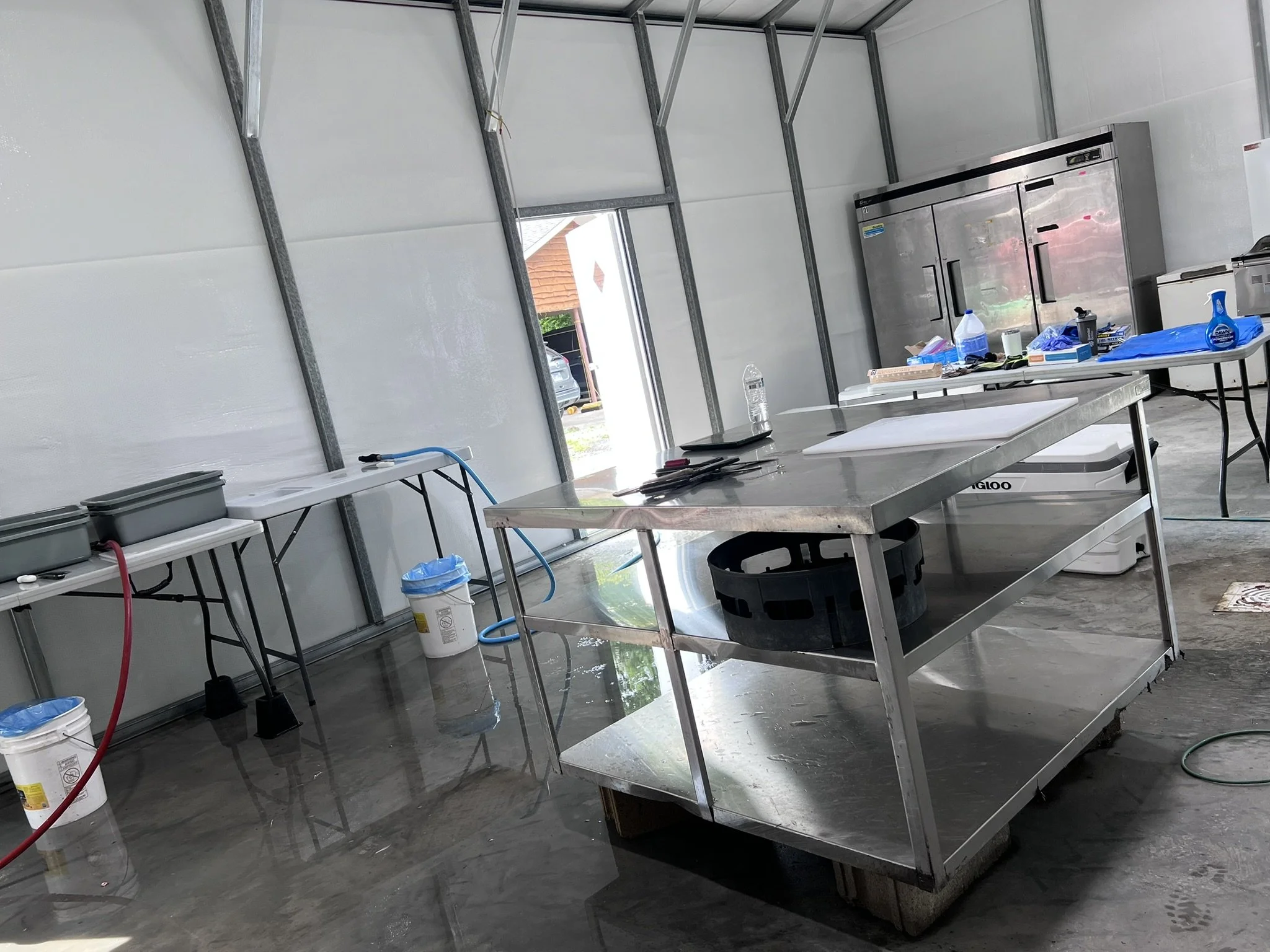 A partly constructed commercial kitchen with metal framing, stainless steel tables, and various cleaning supplies scattered around.