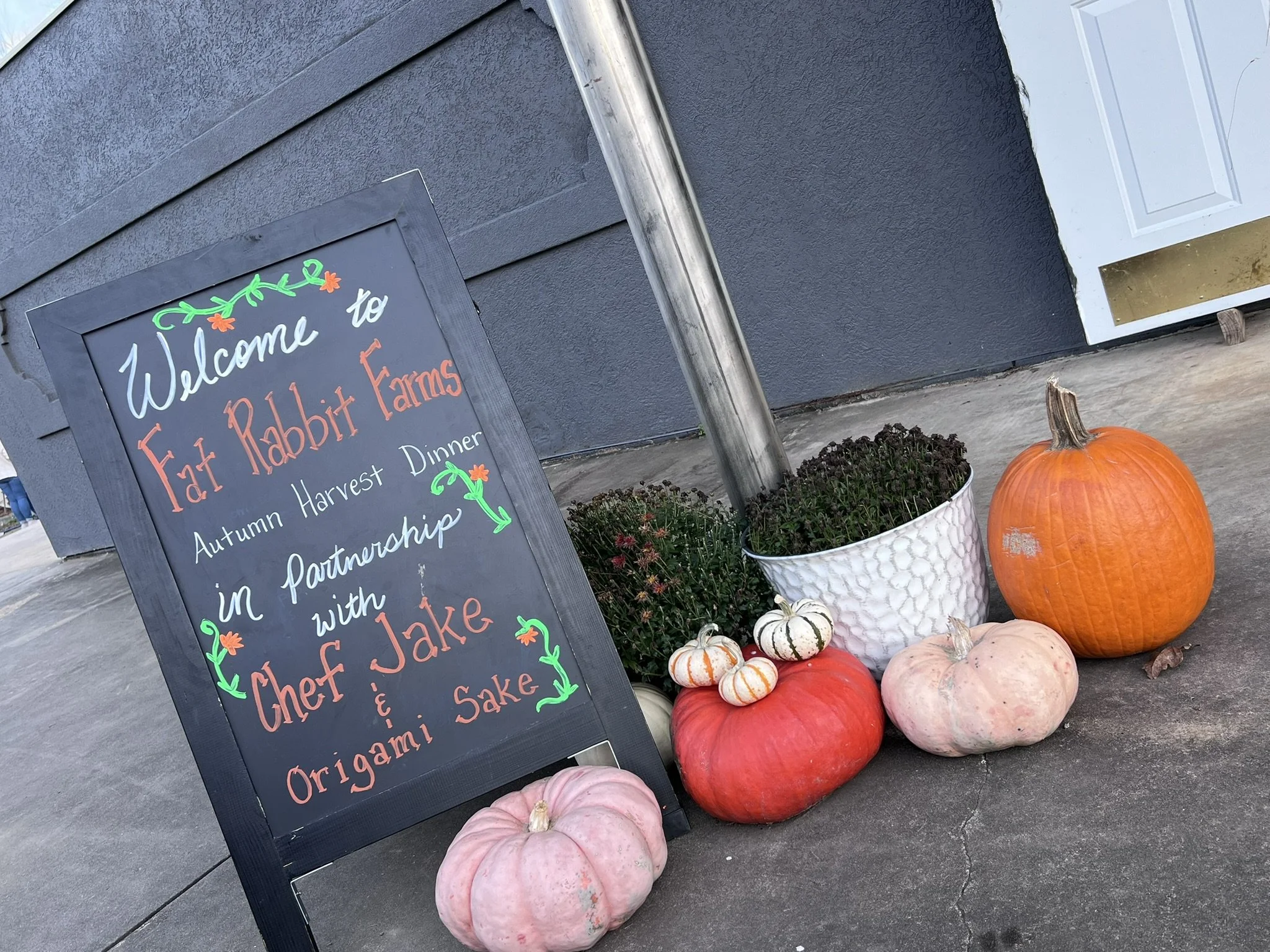 A chalkboard sign with colorful lettering and decorations welcomes visitors to a farm-to-table eatery, 'Fat Rabbit Farms,' highlighting their focus on harvest dinners, autumn harvest partnerships, and homemade dishes.