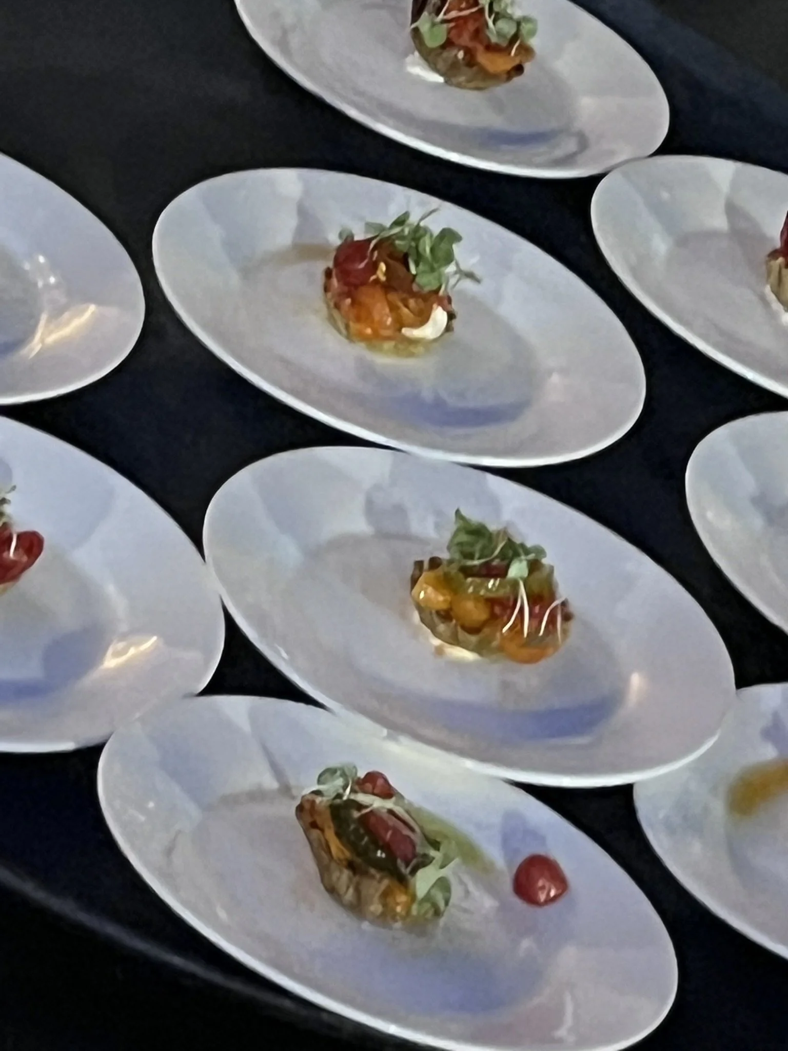 Multiple white oval plates with small servings of food, including a colorful dish garnished with microgreens and a cherry tomato, on a black surface.