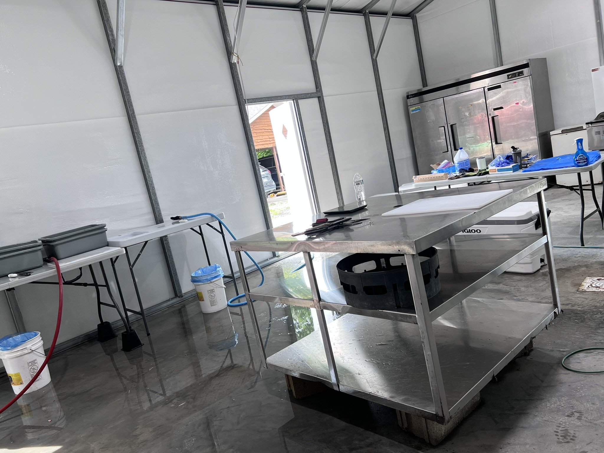 Interior of a utility or workshop space with metal tables, buckets, a water bottle, tools, a large stainless steel appliance, and a partially open door showing a car parked outside.