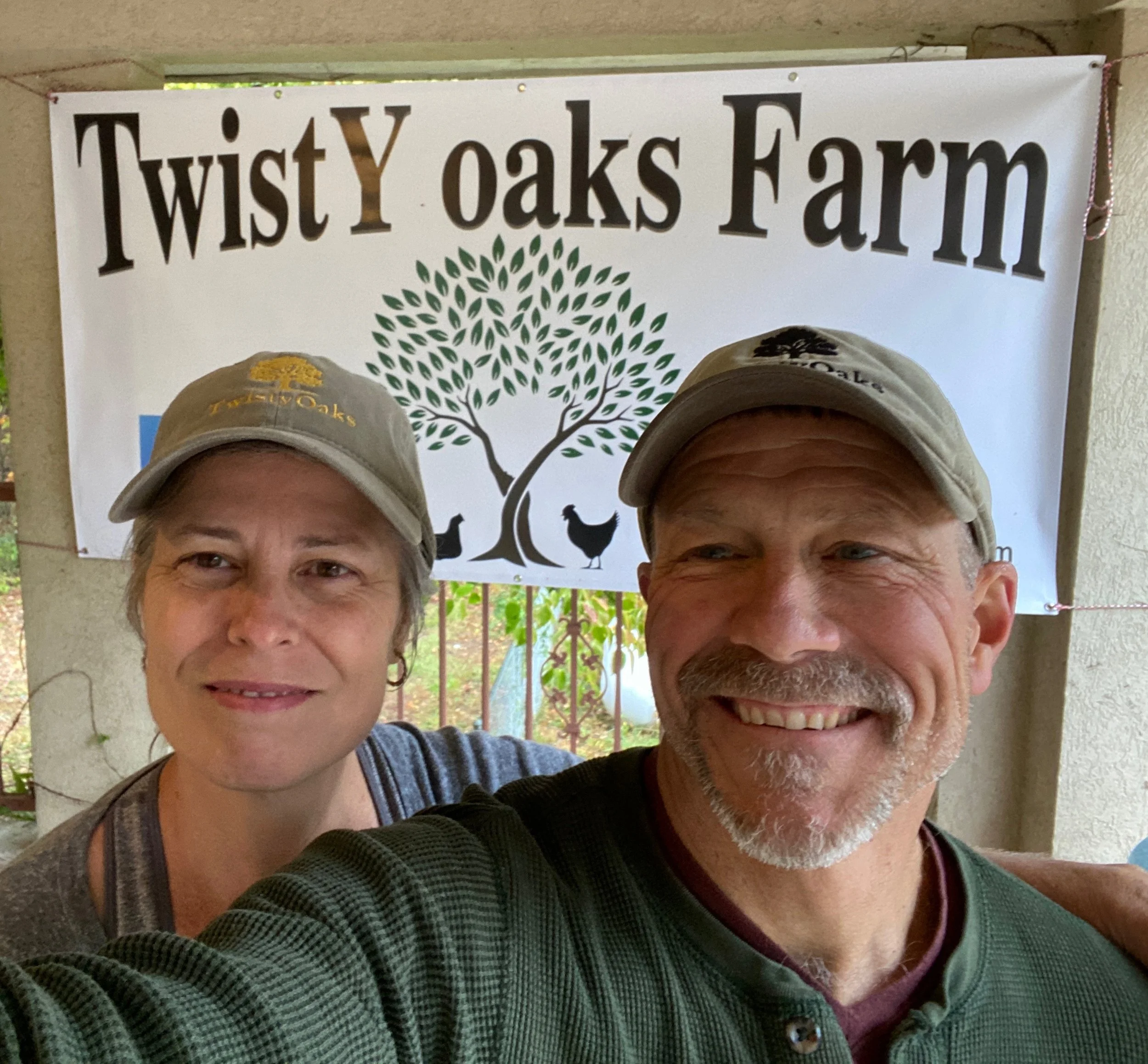 A man and a woman taking a selfie in front of a sign that reads 'Twist Oaks Farm' with a tree graphic and some animal silhouettes underneath. Both are wearing tan caps, and only part of their faces are visible in the photo.