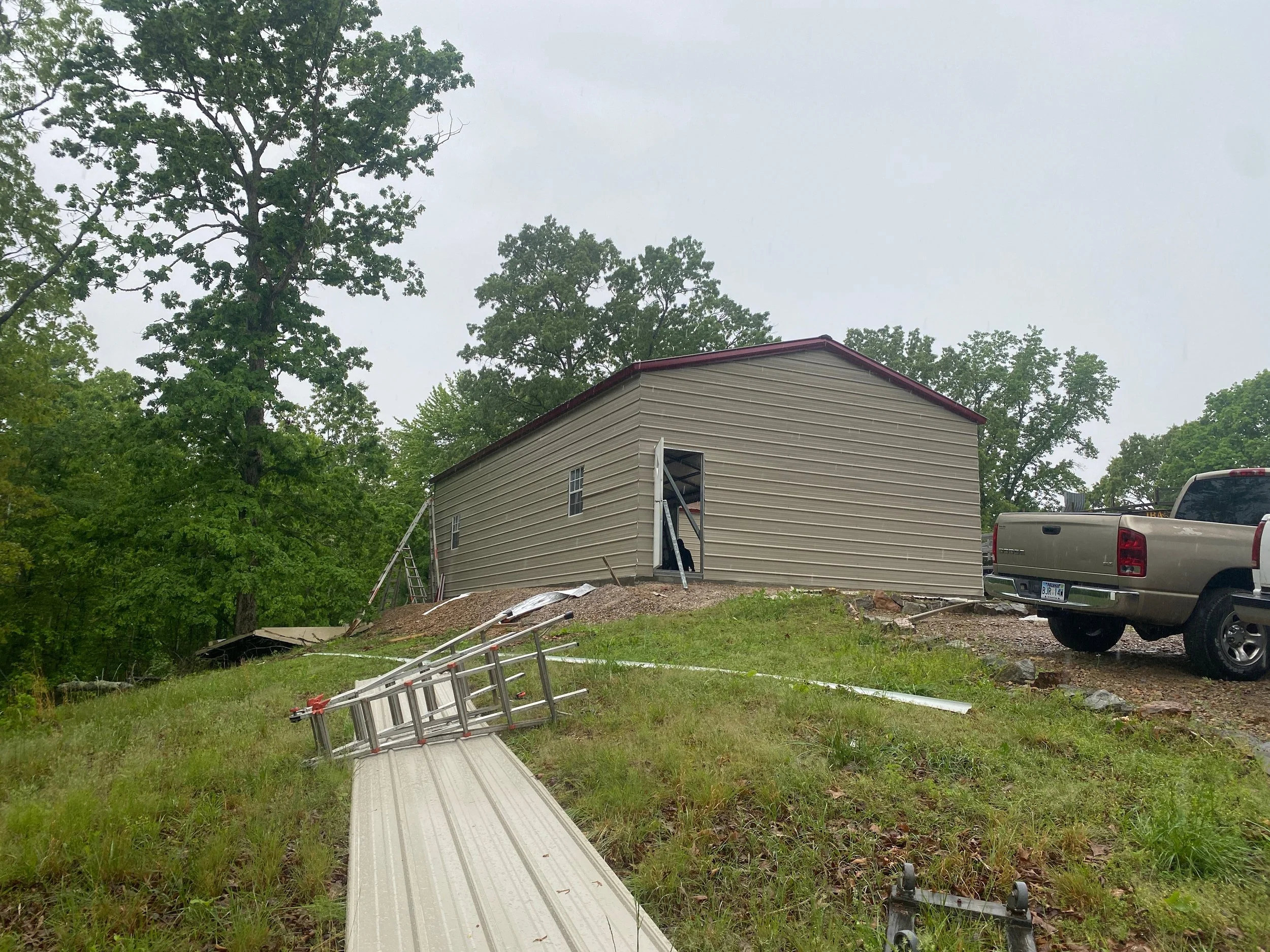 A house being built with siding, stairs, and a metal foundation ramp leading up to the entrance. There are ladders and construction materials on the ground. Several trucks are parked nearby, and trees surround the scene under an overcast sky.