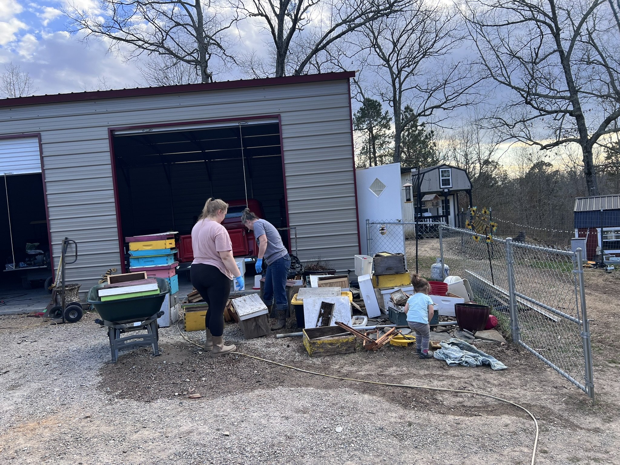 Two women and a young girl sorting through various items and furniture outside a metal storage shed in a backyard. The scene appears to be a yard sale or cleanup event, with scattered objects and storage containers. Background includes trees, a chain