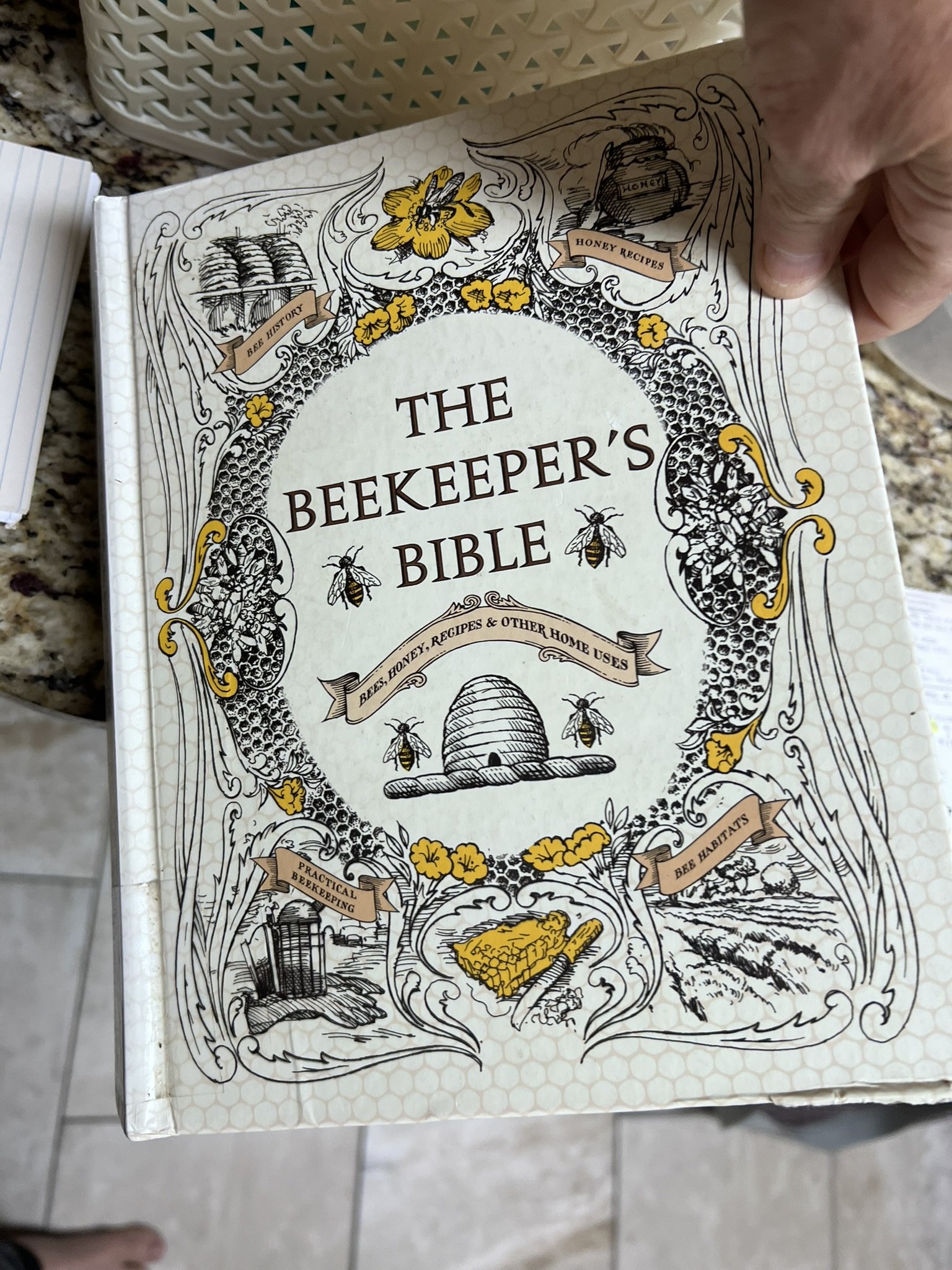 Cover of a book titled "The Beekeeper's Bible" with illustrations of a hive, bees, honey jar labeled "Honey", a bee on a honeycomb, and other beekeeping-related drawings in black and yellow ink.