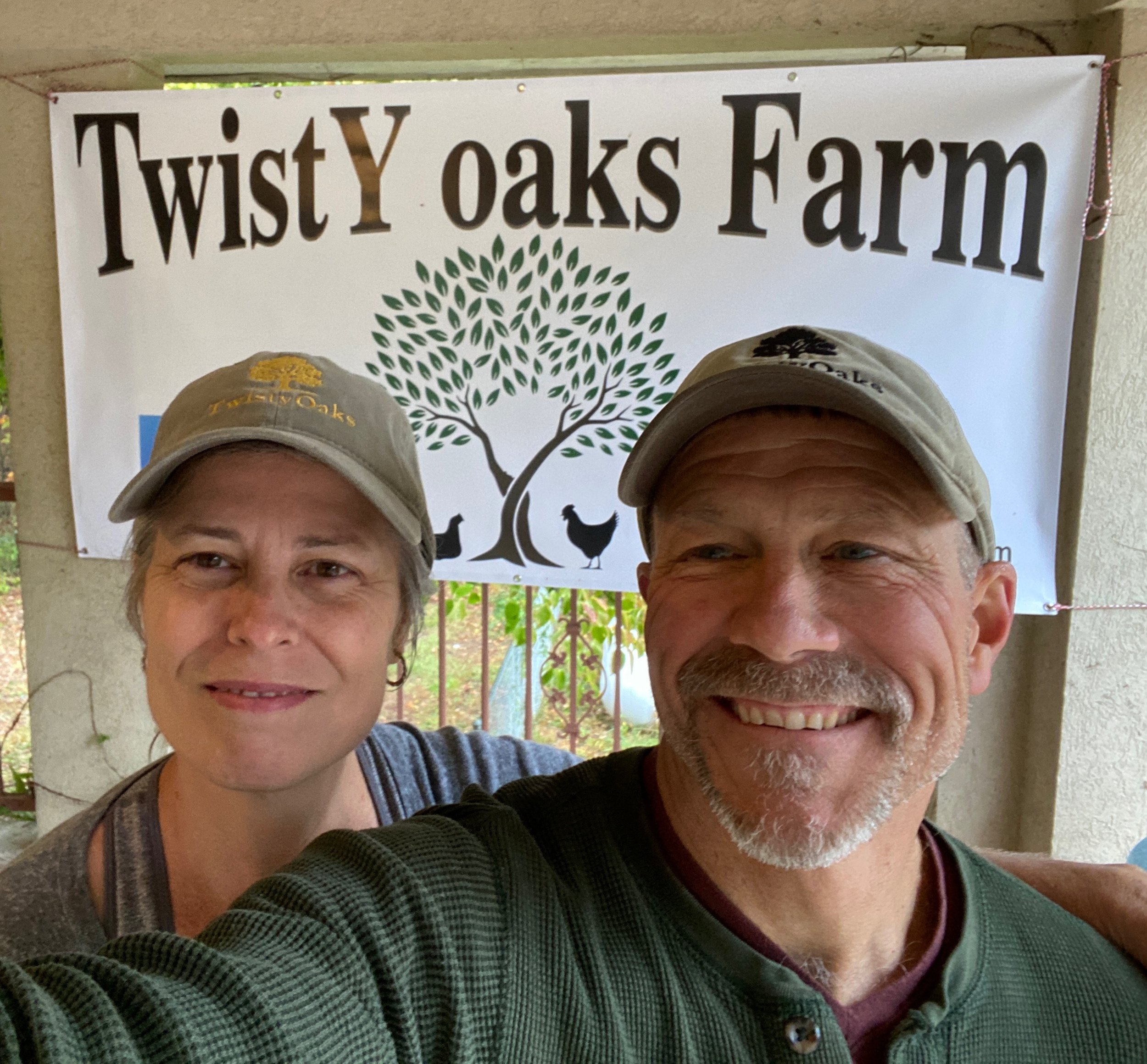 Two people wearing hats stand in front of a sign that reads 'Twist Oaks Farm' with a tree logo above and chickens below.