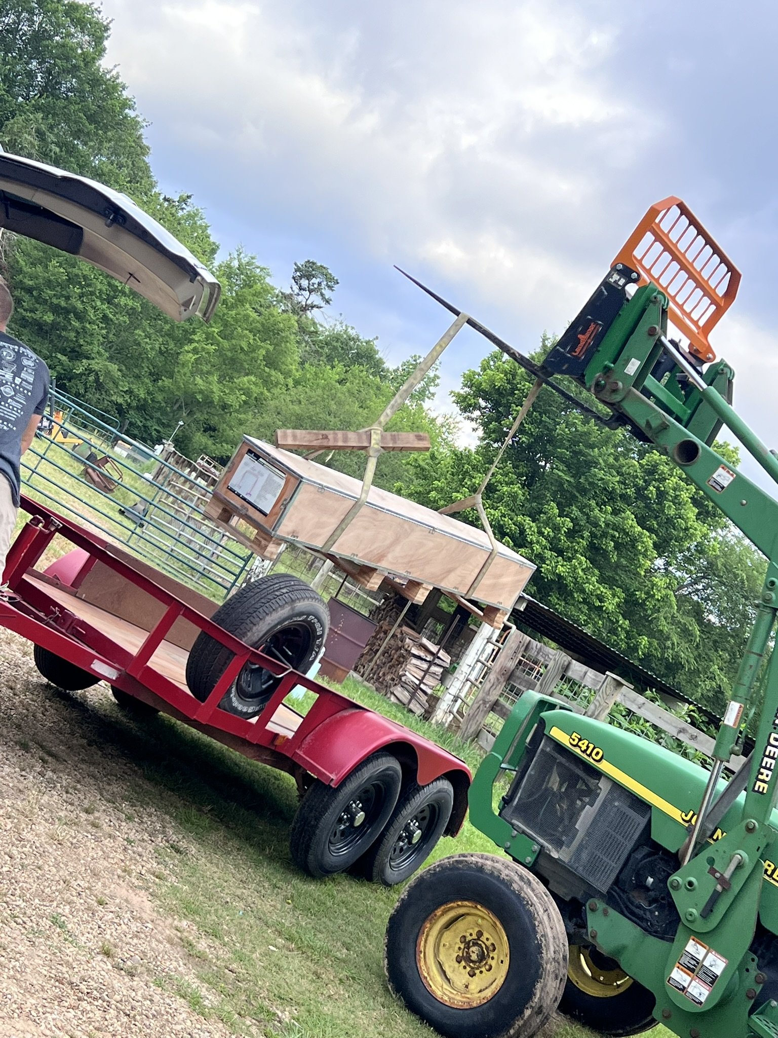 A farm scene with a green John Deere tractor and a pink flatbed trailer attached. The trailer is holding a small wooden structure with wheels, and there are trees and fences in the background under a cloudy sky.