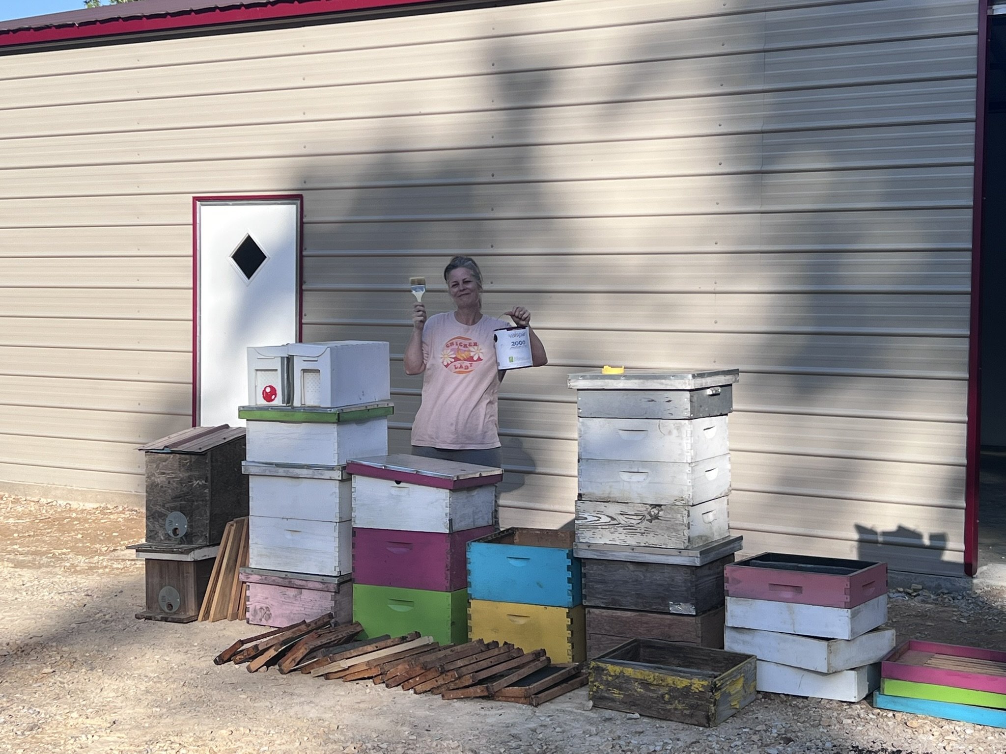 A woman smiling and holding a paintbrush and a container of paint, standing outdoors near a beige metal building with colorful stacked wooden crates and other containers around her.