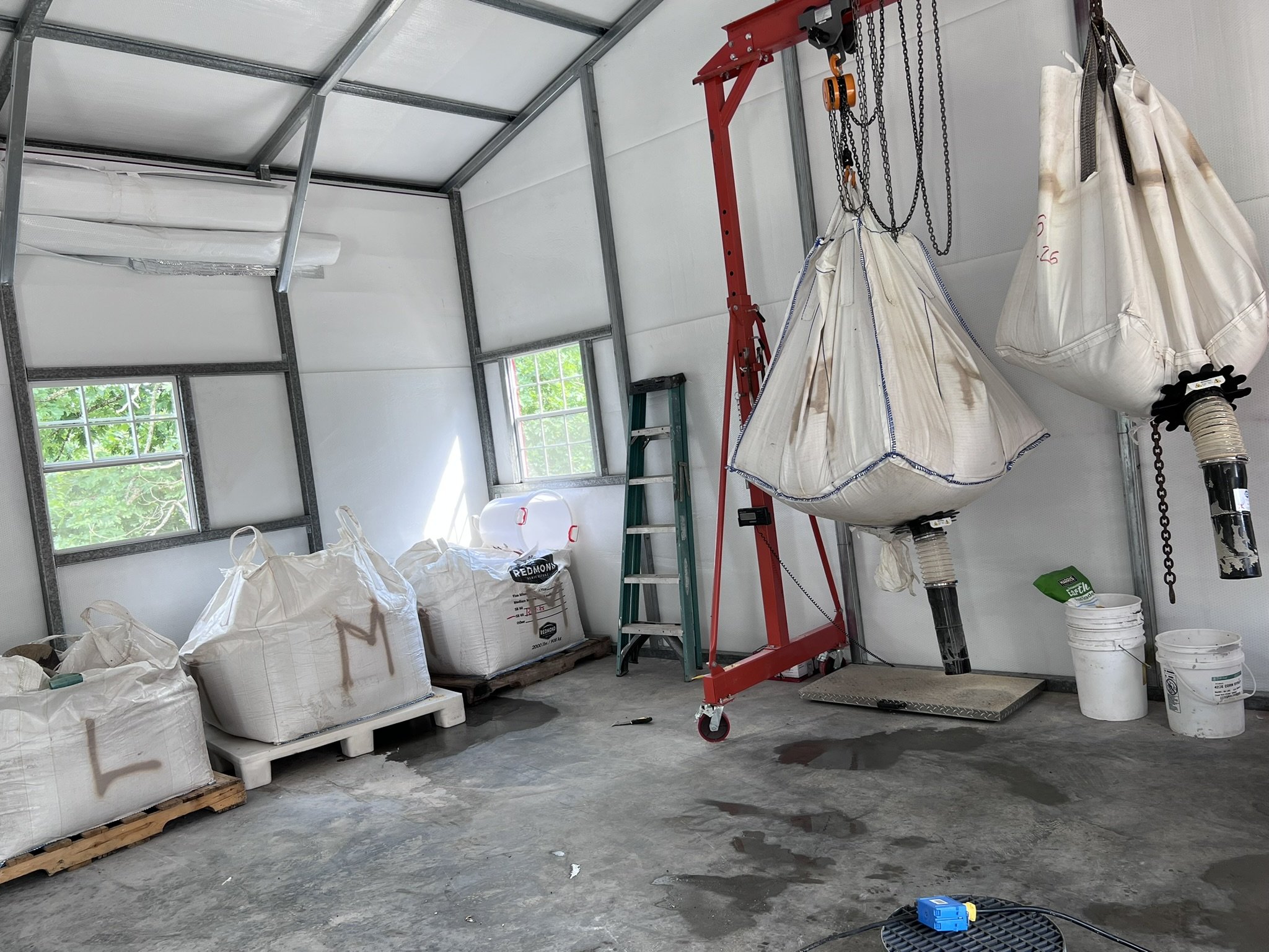 The image shows the interior of a garage under construction with metal framing, a ladder, bags of construction material, and a large hanging fabric structure supported by chains. There are portable windows on the wall and buckets on the floor.