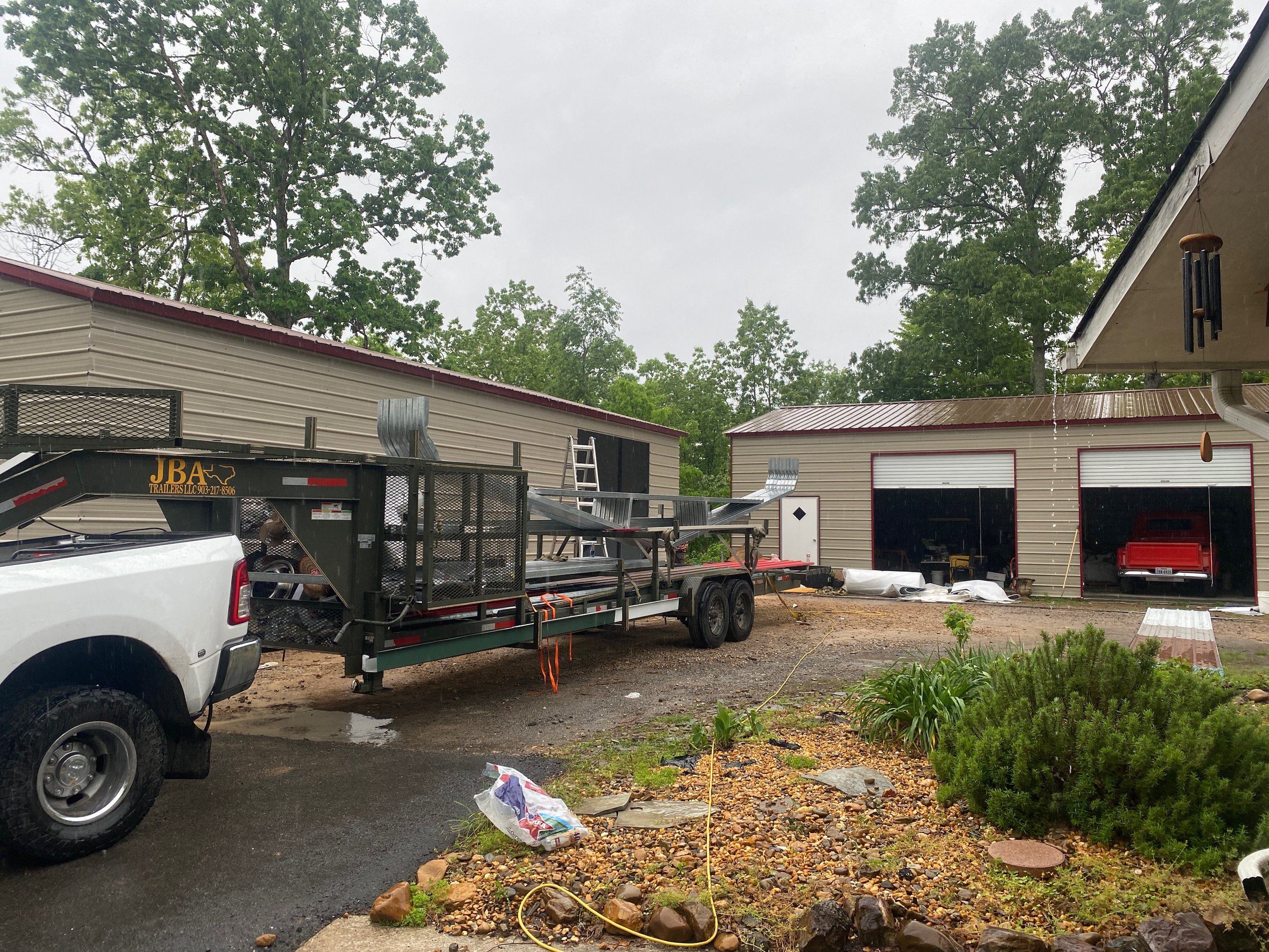 A white pickup truck and utility trailers parked in front of a beige building with red trim. Trees and cloudy sky in the background. Items on the ground include a plastic bag, rocks, and a garden hose.