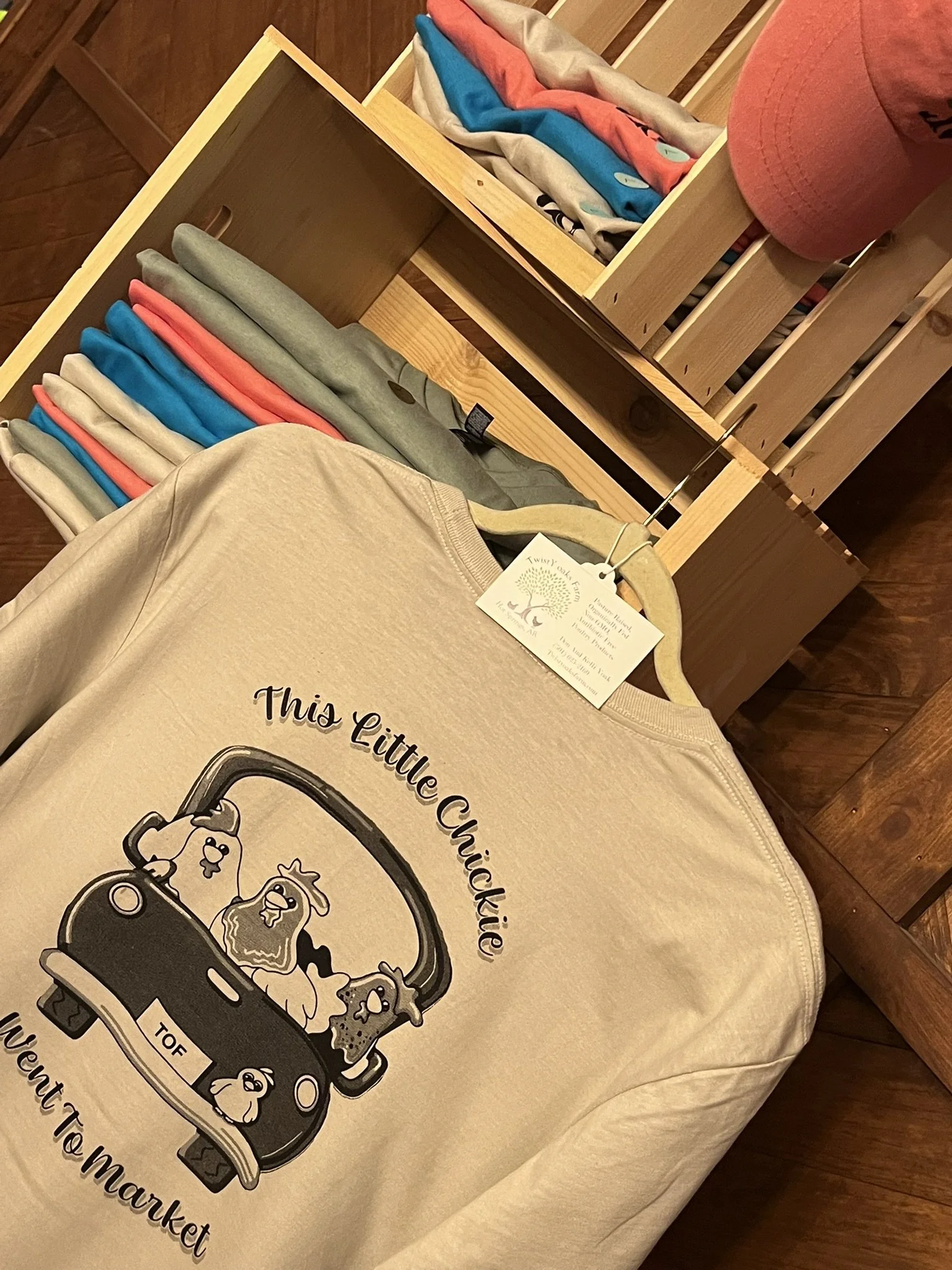 A beige T-shirt on a wooden table with a graphic of chickens in a market truck and the text 'This Little Chicken'.