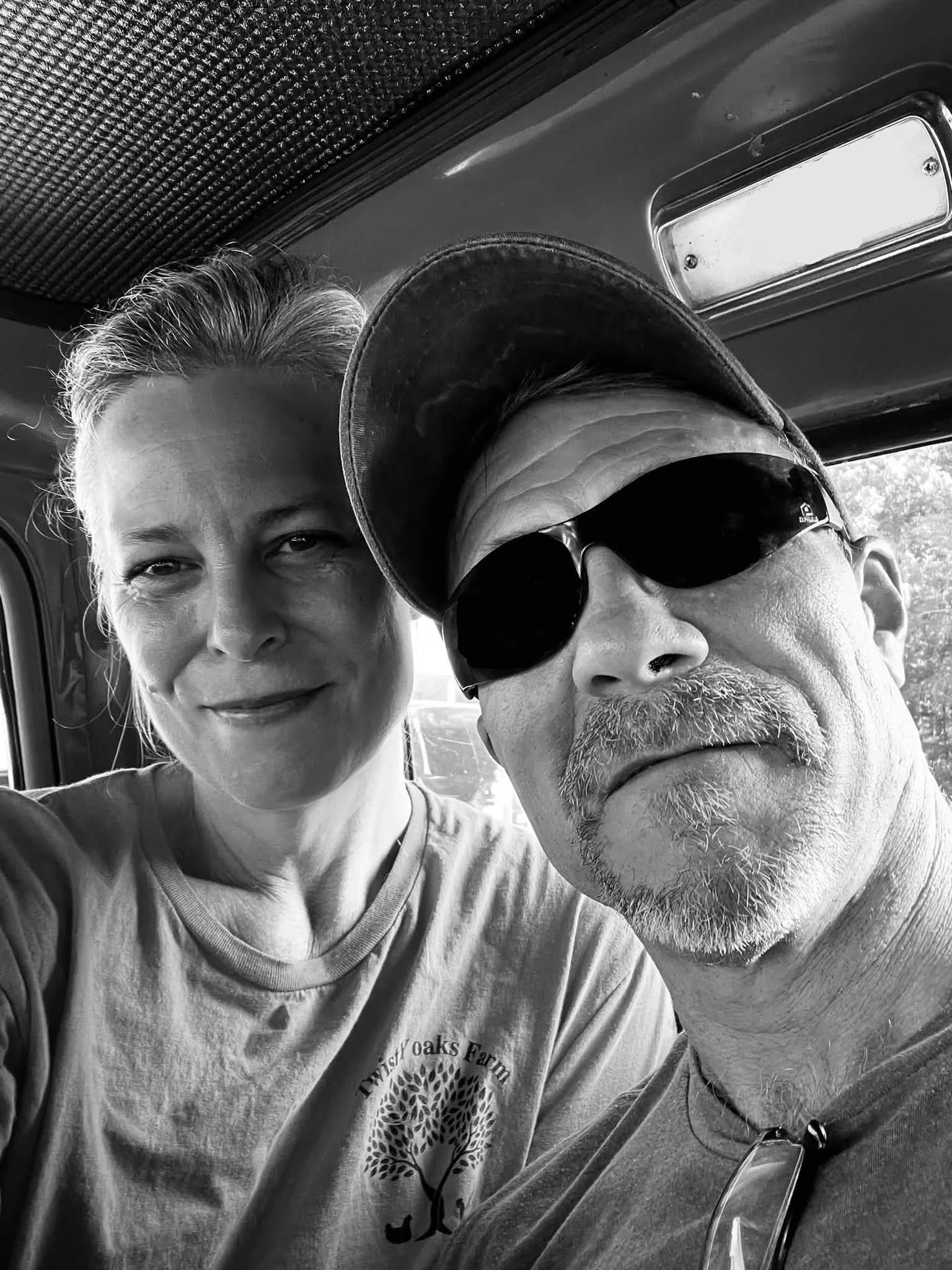 A black and white photo of two people taking a close-up selfie inside a vehicle. The woman on the left has short, light-colored hair and is smiling at the camera. The man on the right is wearing a baseball cap, sunglasses, and has a beard and mustache.