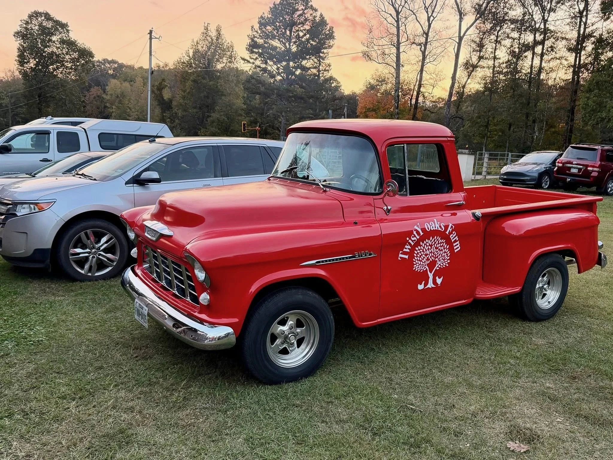 A red vintage Chevrolet pickup truck with a farm logo on the door parked on the grass, surrounded by modern cars, with trees and a sunset in the background.