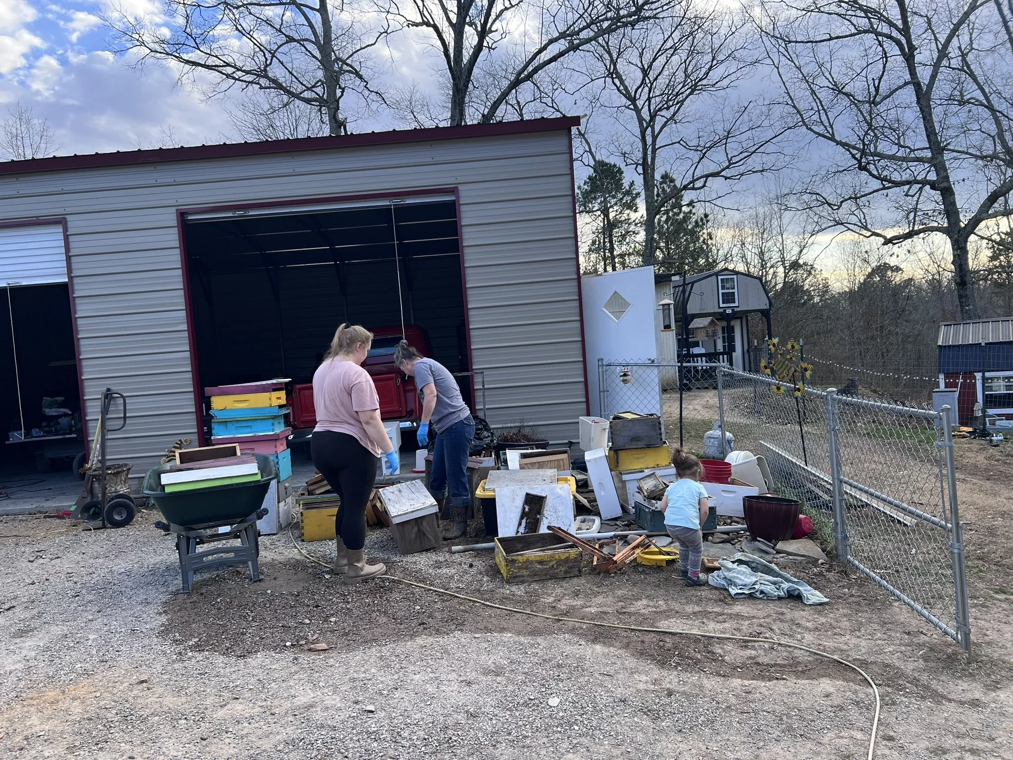 People organizing and sorting items outdoors near a metal shed, with various containers, furniture, and garden decorations around them, and trees in the background.