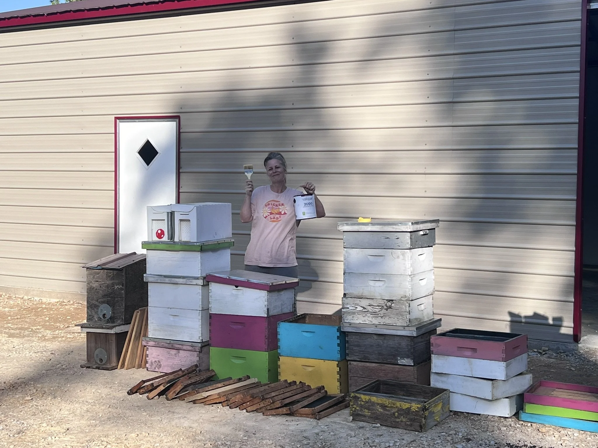 A woman standing outdoors next to colorful stacked wooden crates and boxes, holding a paintbrush and a container of paint, with a beige metal building in the background.