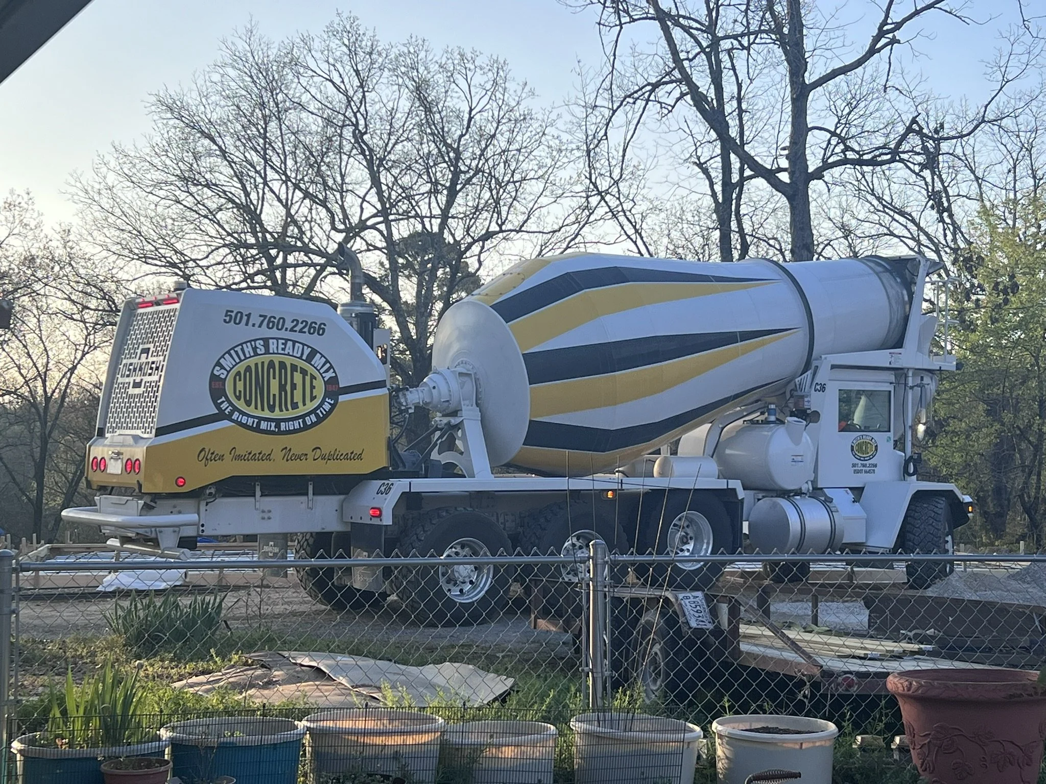 A cement mixer truck with white, yellow, and black colors parked behind a chain-link fence in a yard with potted plants and trees with no leaves.