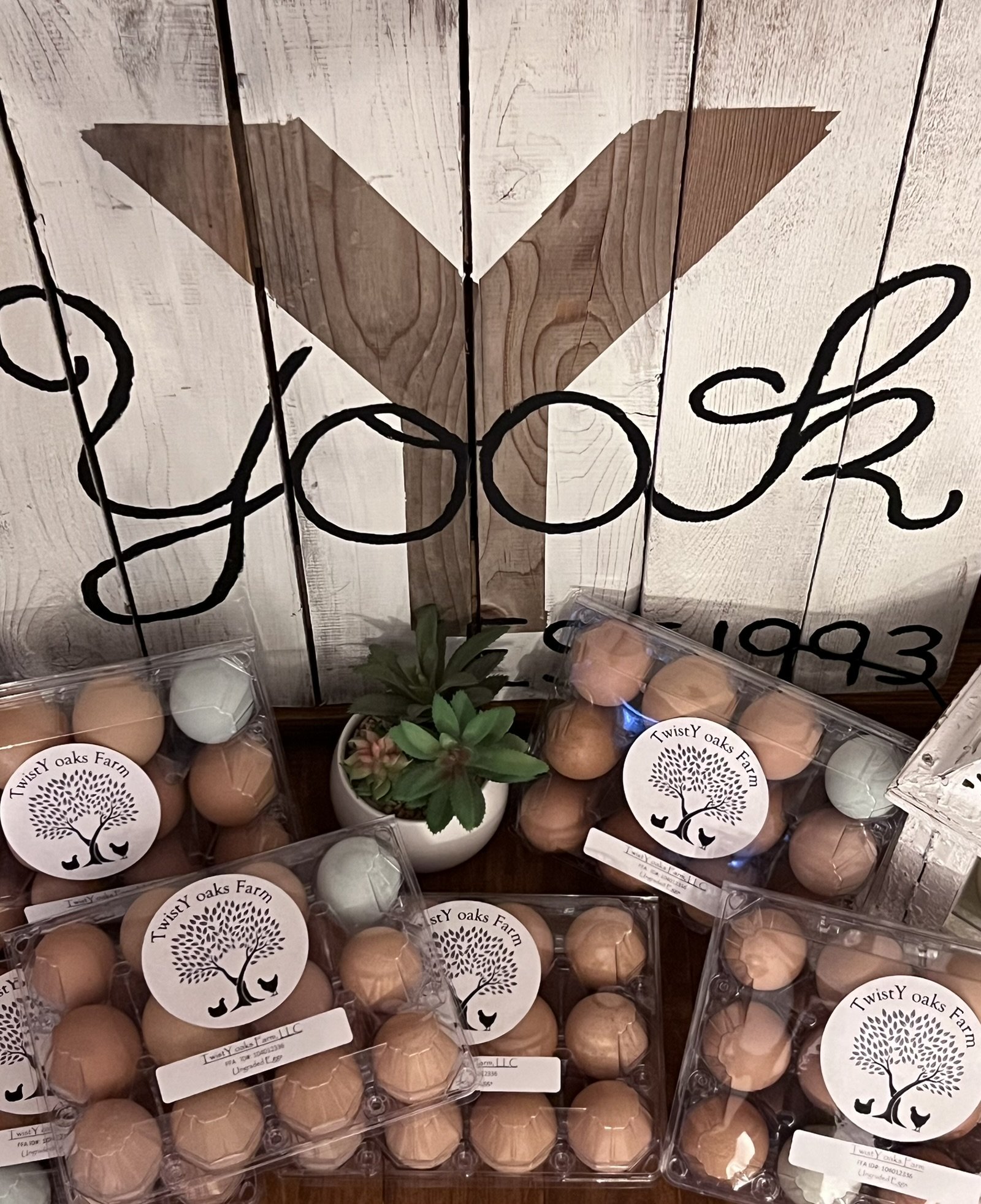 Eggs in clear cartons with 'TwistY oaks Farm' stickers, a small plant, and a wooden wall with black writing.