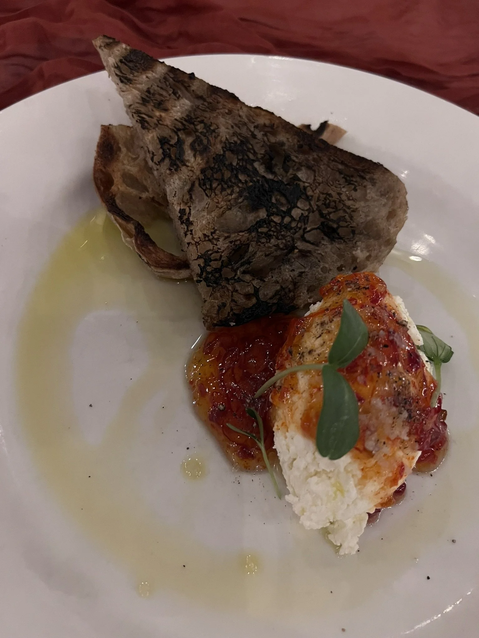 A piece of grilled fish with crispy skin served on a white plate with a small dollop of tomato-based sauce, garnished with fresh herbs, and a drizzle of oil or sauce.