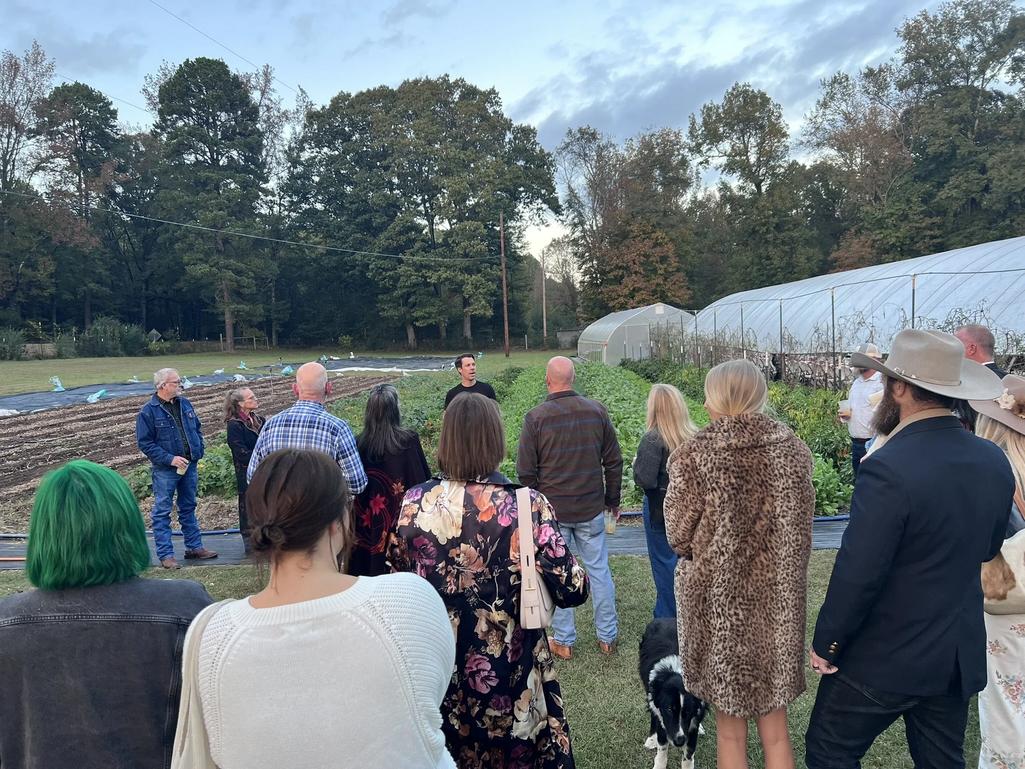 A group of people gathered outdoors near greenhouses, listening to a man speaking. The group includes men and women, some with brightly colored hair. The setting is a farm or garden with trees and sky in the background.
