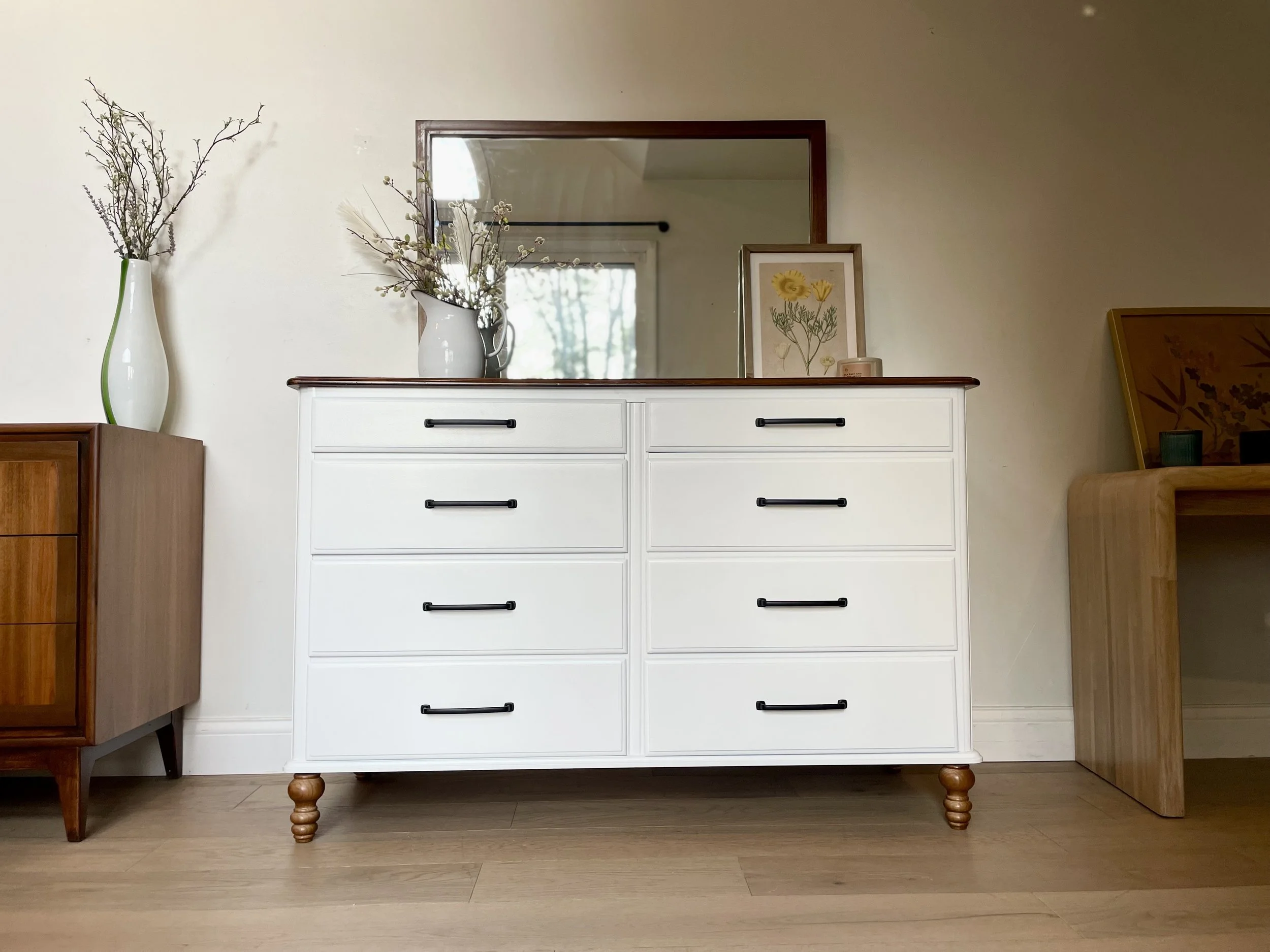 White dresser with black handles and wooden legs, decorated with vases and framed artwork, on a light wood floor against a cream wall.
