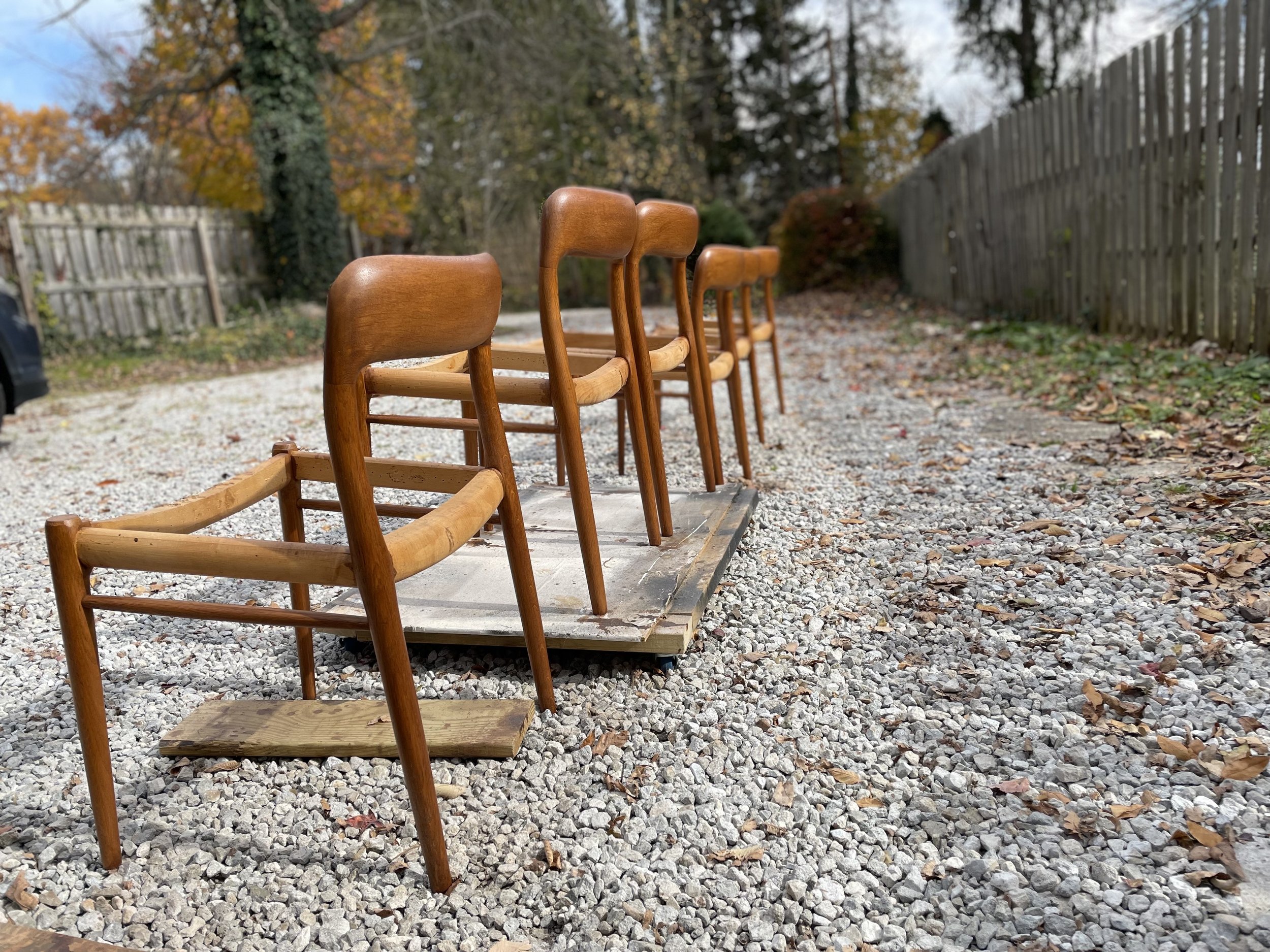 Four wooden chairs are lined up on a gravel surface outdoors, with a wooden plank beneath them. There is a wooden fence on the right and some trees with autumn leaves in the background.