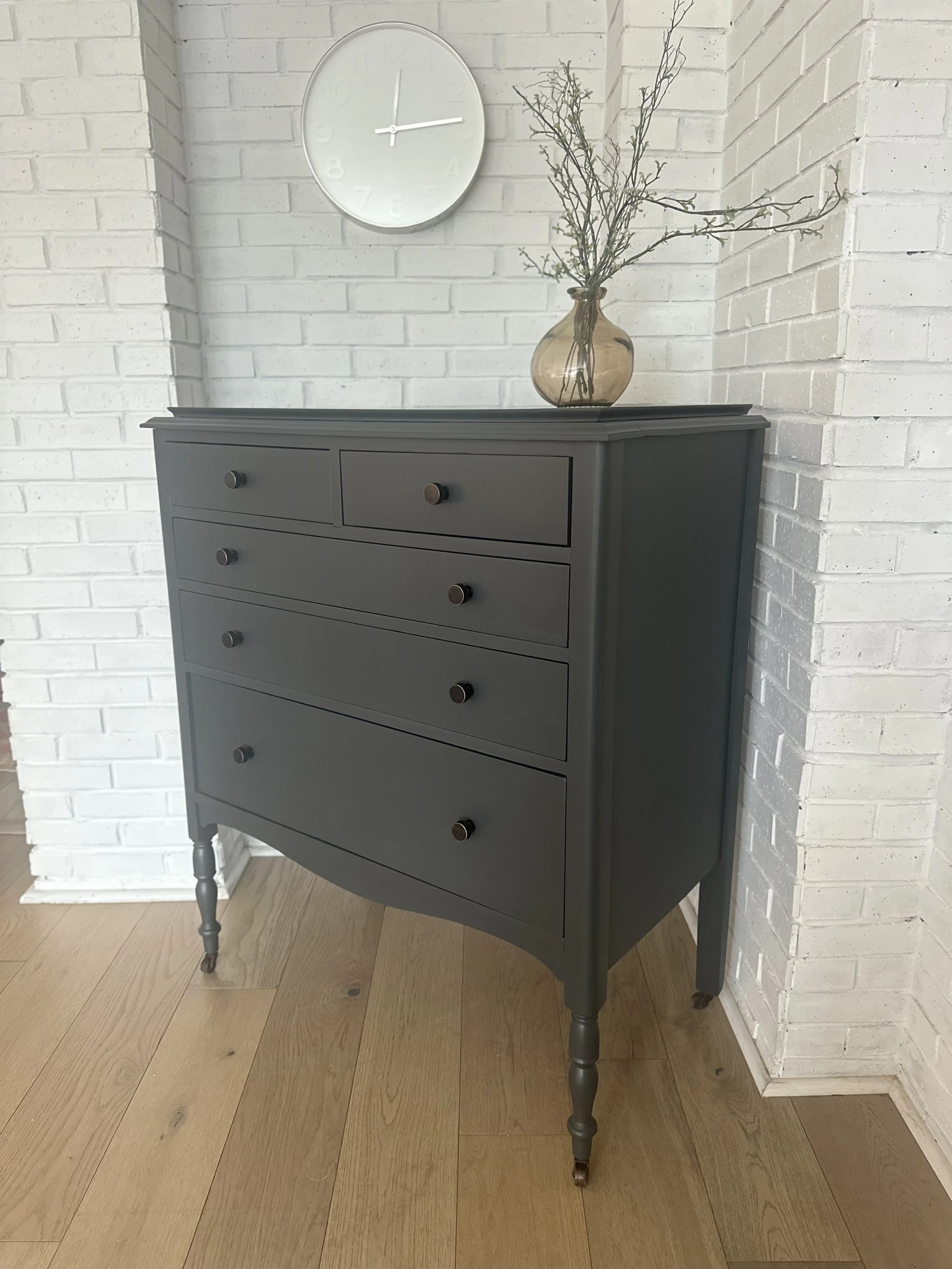 A black dresser with multiple drawers and wooden knobs. On top, there's a amber glass vase with thin branches. A white wall clock showing the time as approximately 2:15 hangs on the white brick wall above the dresser. The floor is wooden.