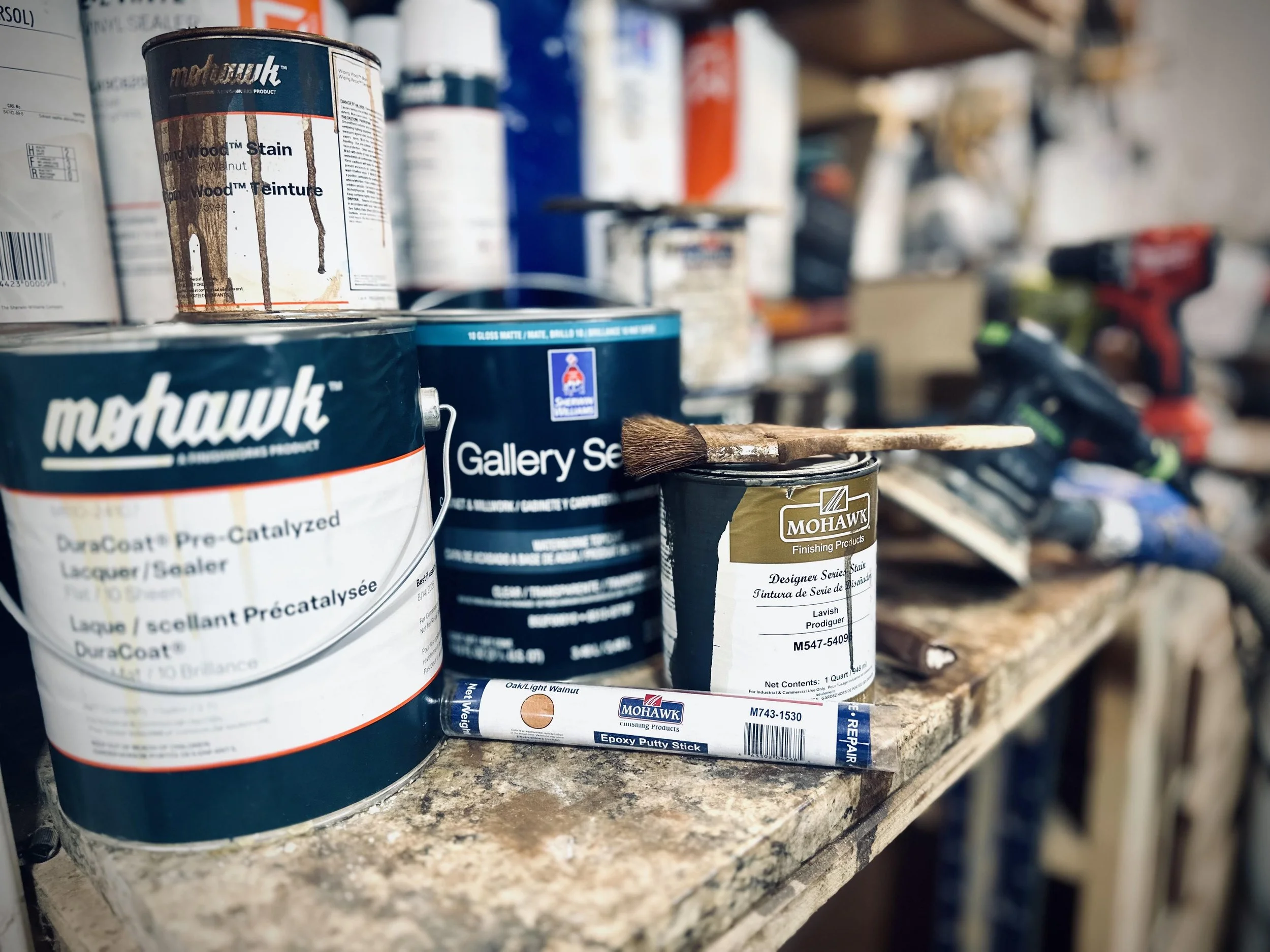 A workbench with cans of wood stain, a paintbrush, a glue stick, and power tools in a workshop.
