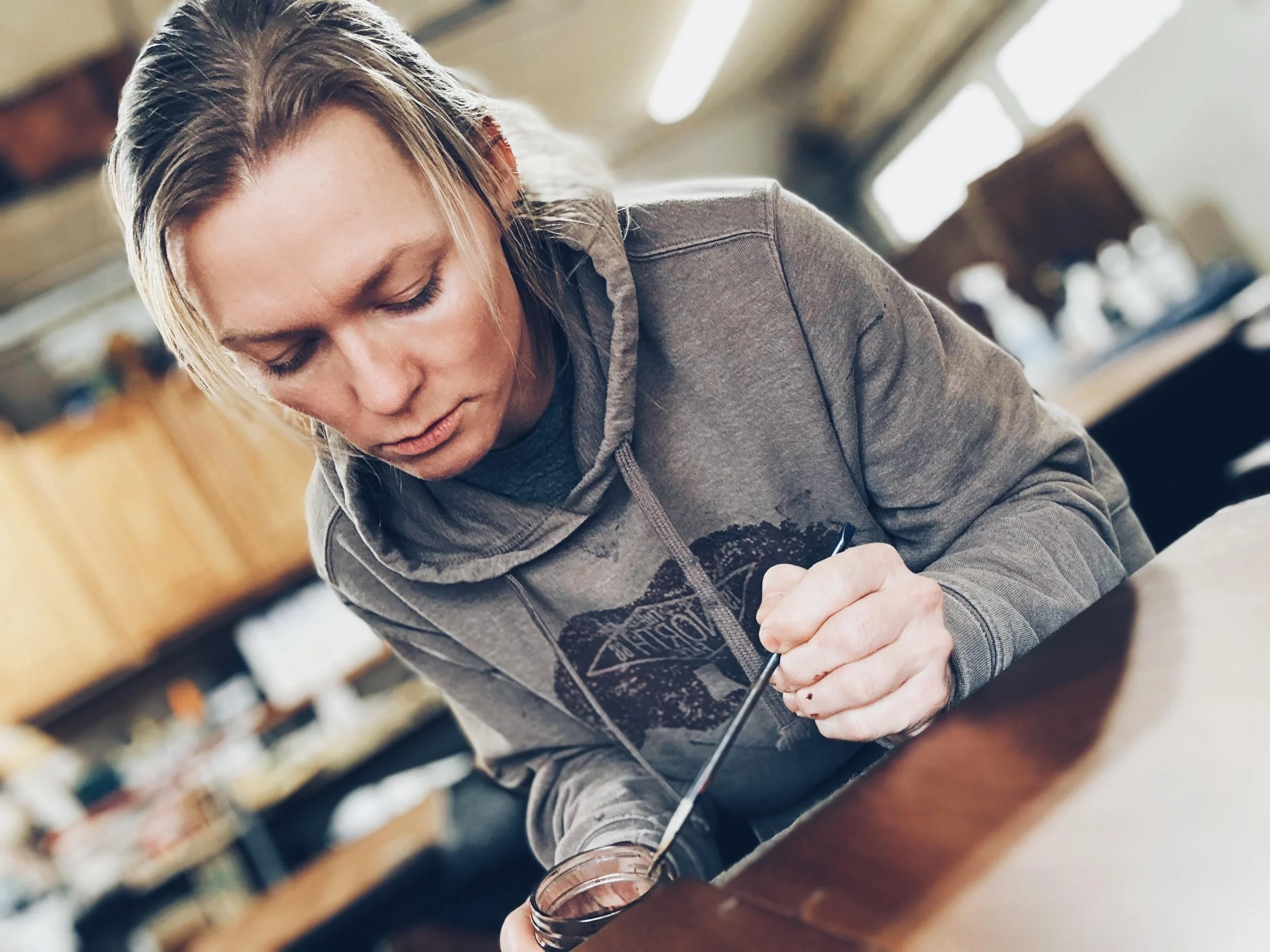 A woman with blonde hair in a gray hoodie decorating a cake with a small paintbrush in a kitchen or bakery setting.