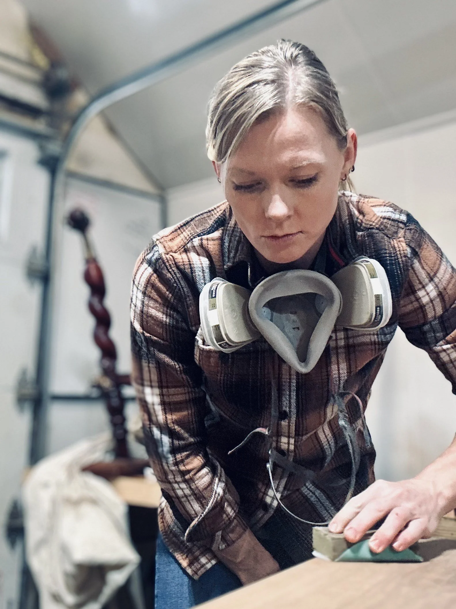 A woman wearing a plaid shirt and a respirator mask on her neck is sanding a piece of wood with a sanding block in a workshop.