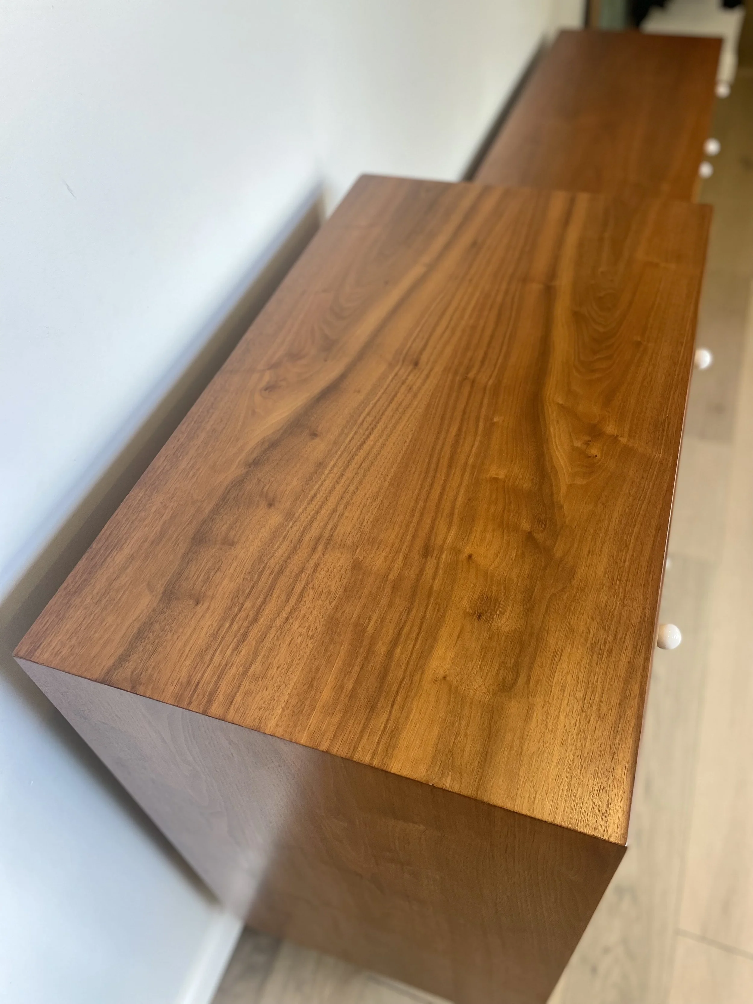 Close-up of a wooden dresser with a smooth, polished surface and white knobs on the side, placed against a wall.