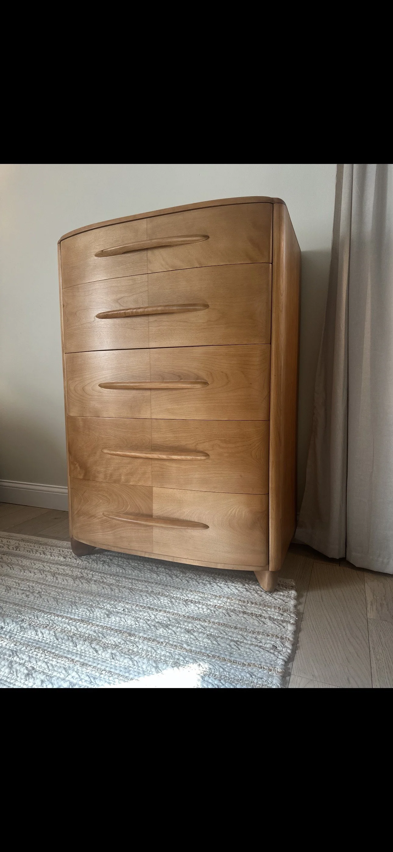 A wooden chest of drawers with five curved drawers and rounded edges, positioned on a patterned rug beside a white wall and a curtain.