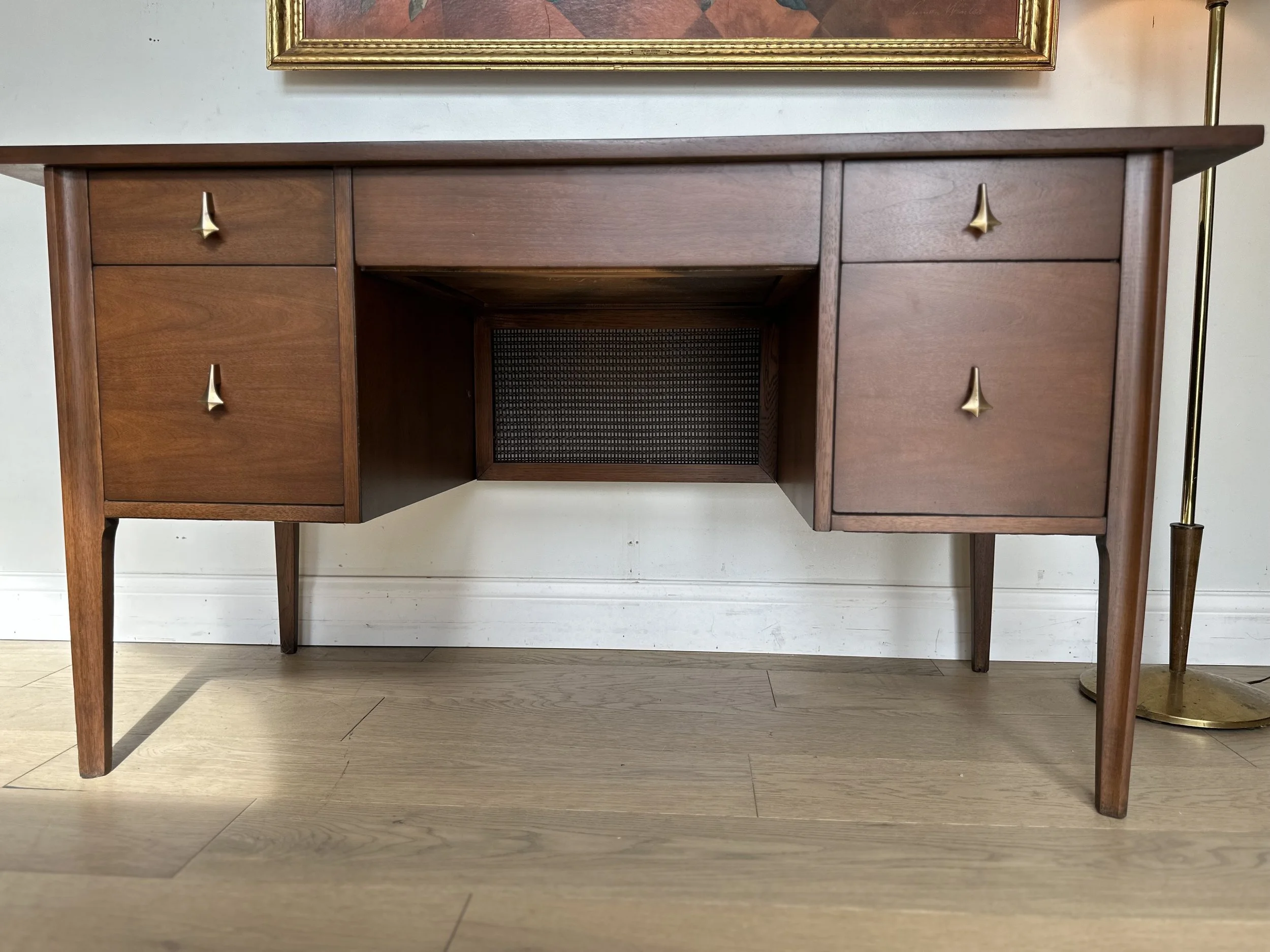 Mid-century modern wooden desk with four drawers, two on each side, and a central open cubby, positioned against a wall with a picture hung above it and a brass floor lamp to the right.