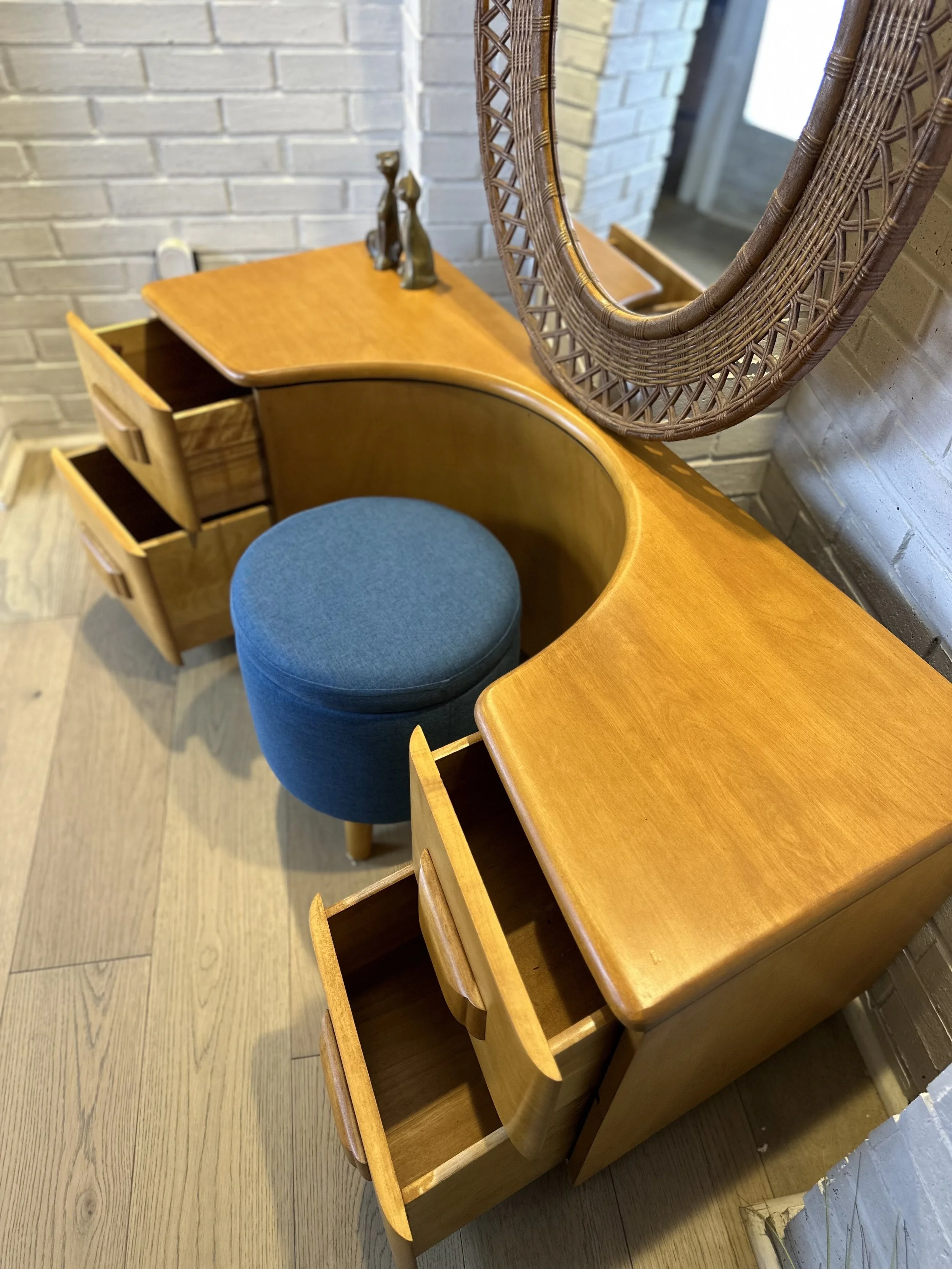A wooden vanity table with drawers, a round blue cushioned stool, a mirror with a cane frame, and decorative figures on top, set against a white brick wall.