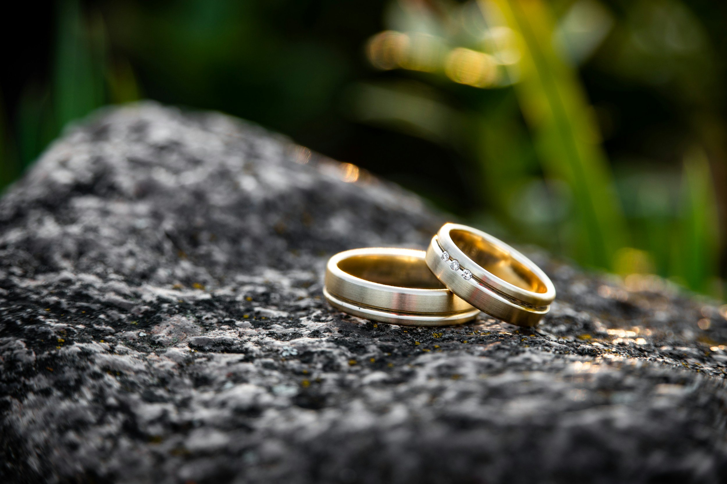 Two wedding rings, one with diamonds, resting on a textured rock surface with green blurred foliage in the background.