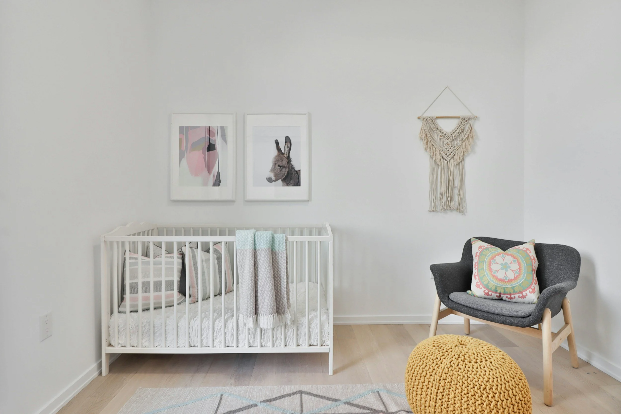 A nursery with a white crib, wall art featuring a colorful cow and a donkey, a beige macrame wall hanging, a gray armchair with a decorative pillow, a large mustard-yellow knitted pouf, and a light-colored wooden floor.