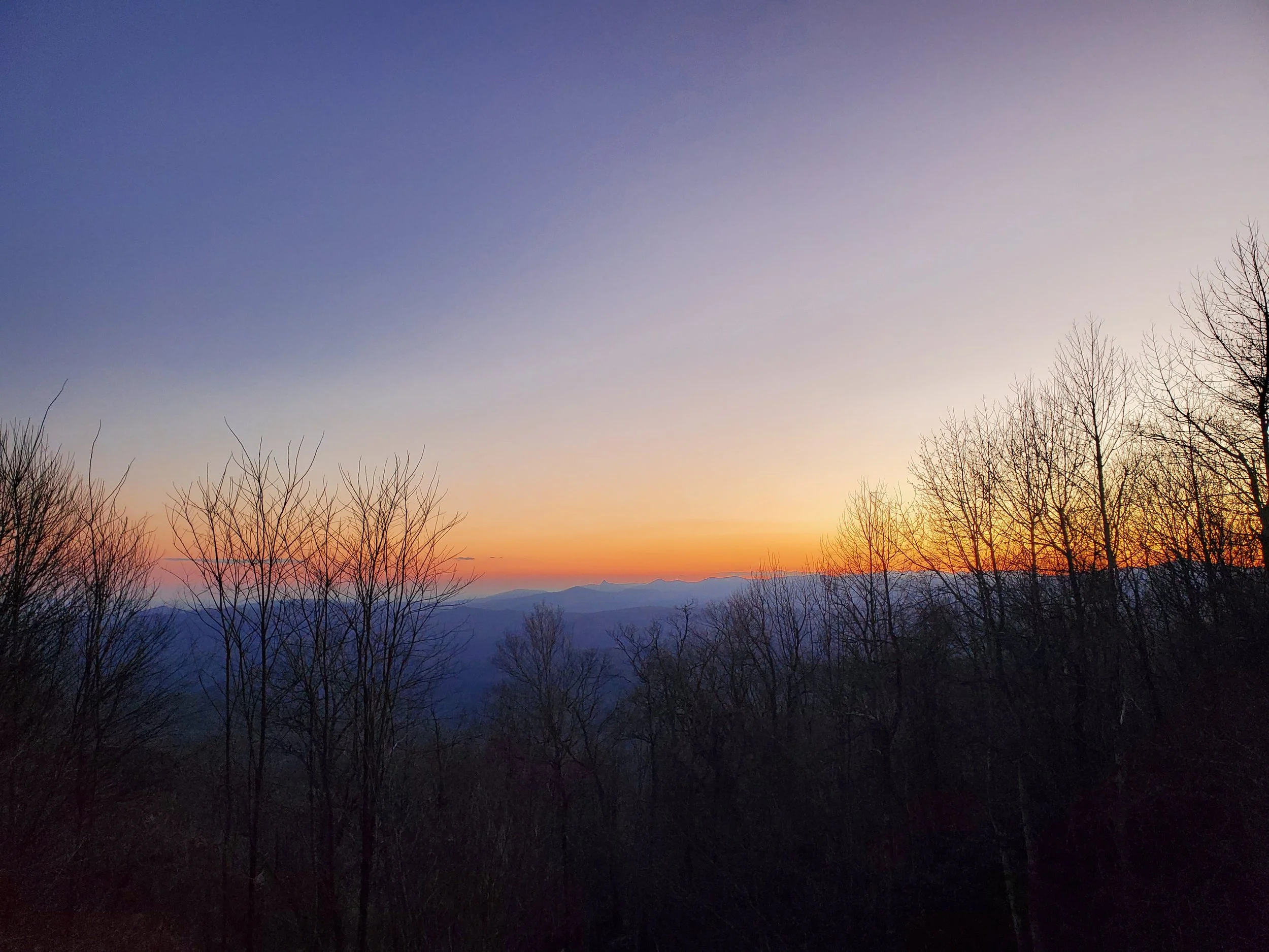 A scenic view of a sunset sky with shades of purple, pink, and orange, and silhouettes of leafless trees in the foreground, with mountains in the distance.