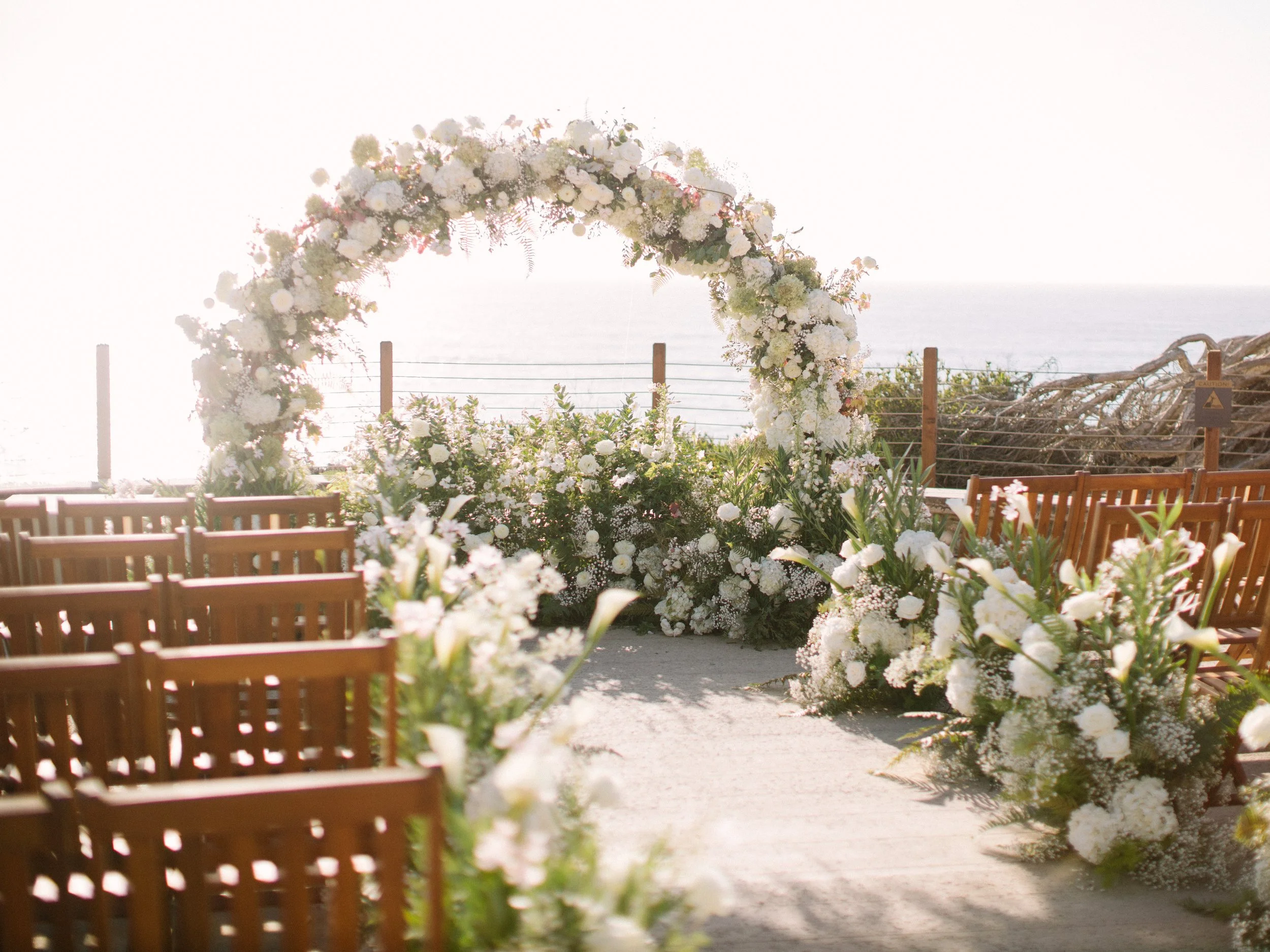 Outdoor wedding altar decorated with white flowers, arch, wooden chairs, and ocean view in the background.