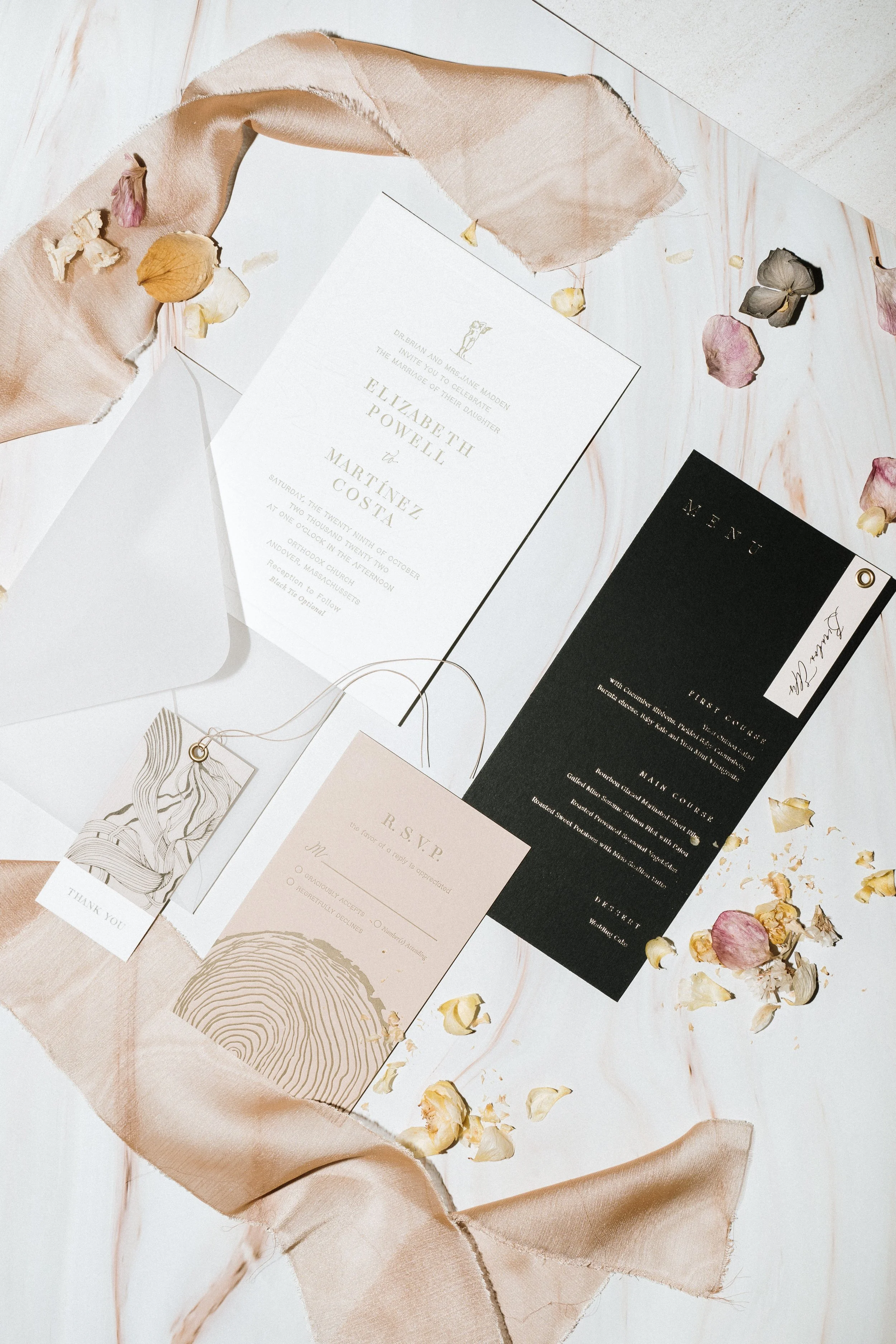 Wedding invitation suite with RSVP card, thank you tag, and menu, surrounded by scattered dried flower petals and a satin ribbon on a white marble surface.