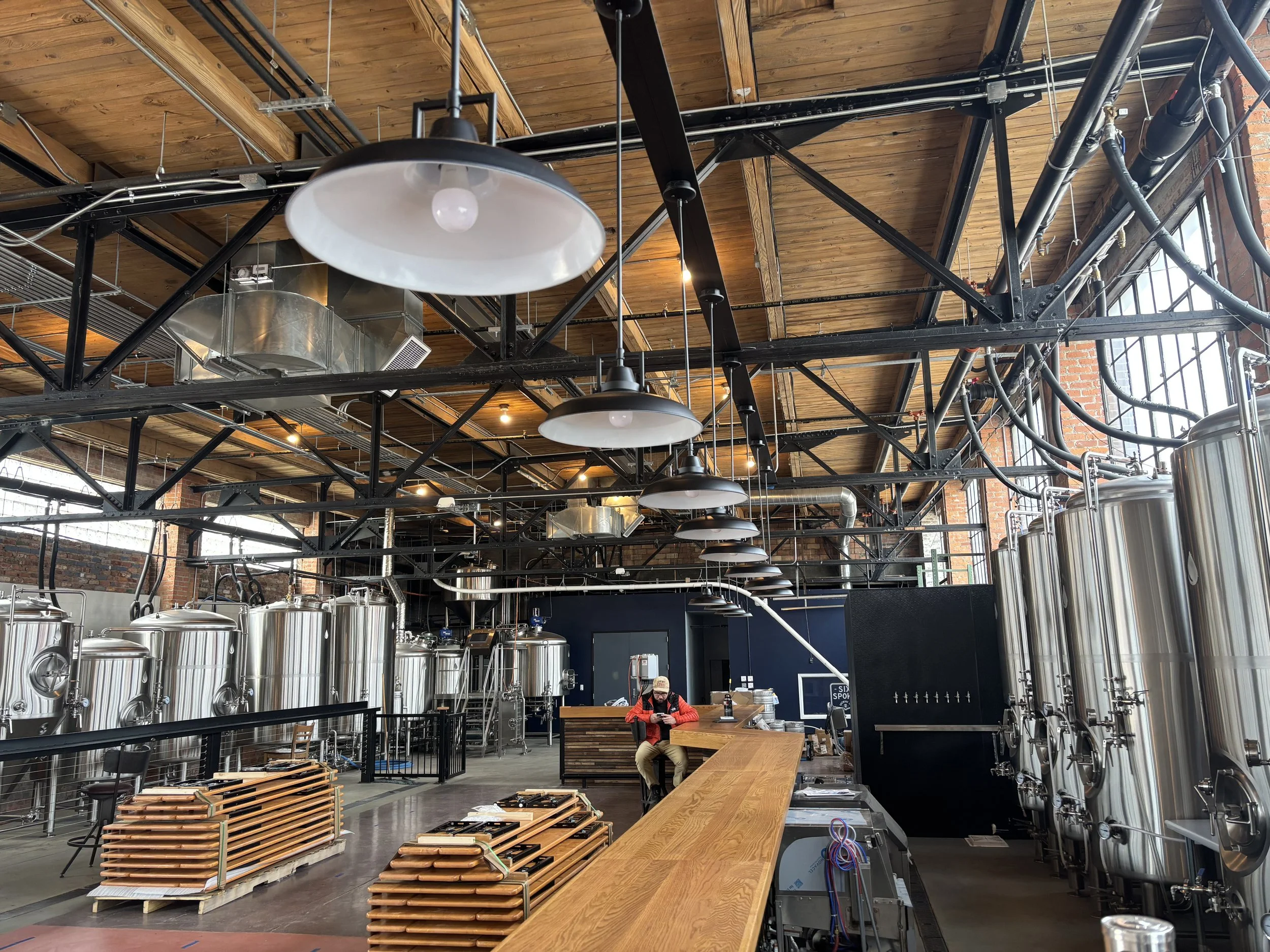 Interior of a brewery with large stainless steel fermentation tanks, wooden ceiling, and a bar with a person sitting and using a phone.