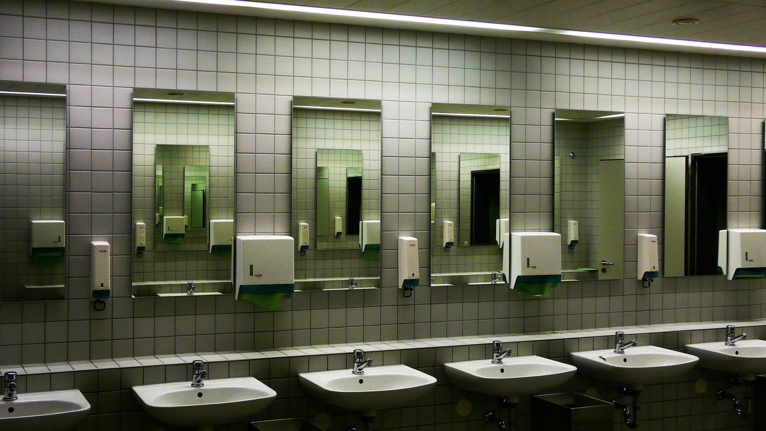 A public restroom with six sinks, six mirrors, and paper towel dispensers on tiled gray walls.