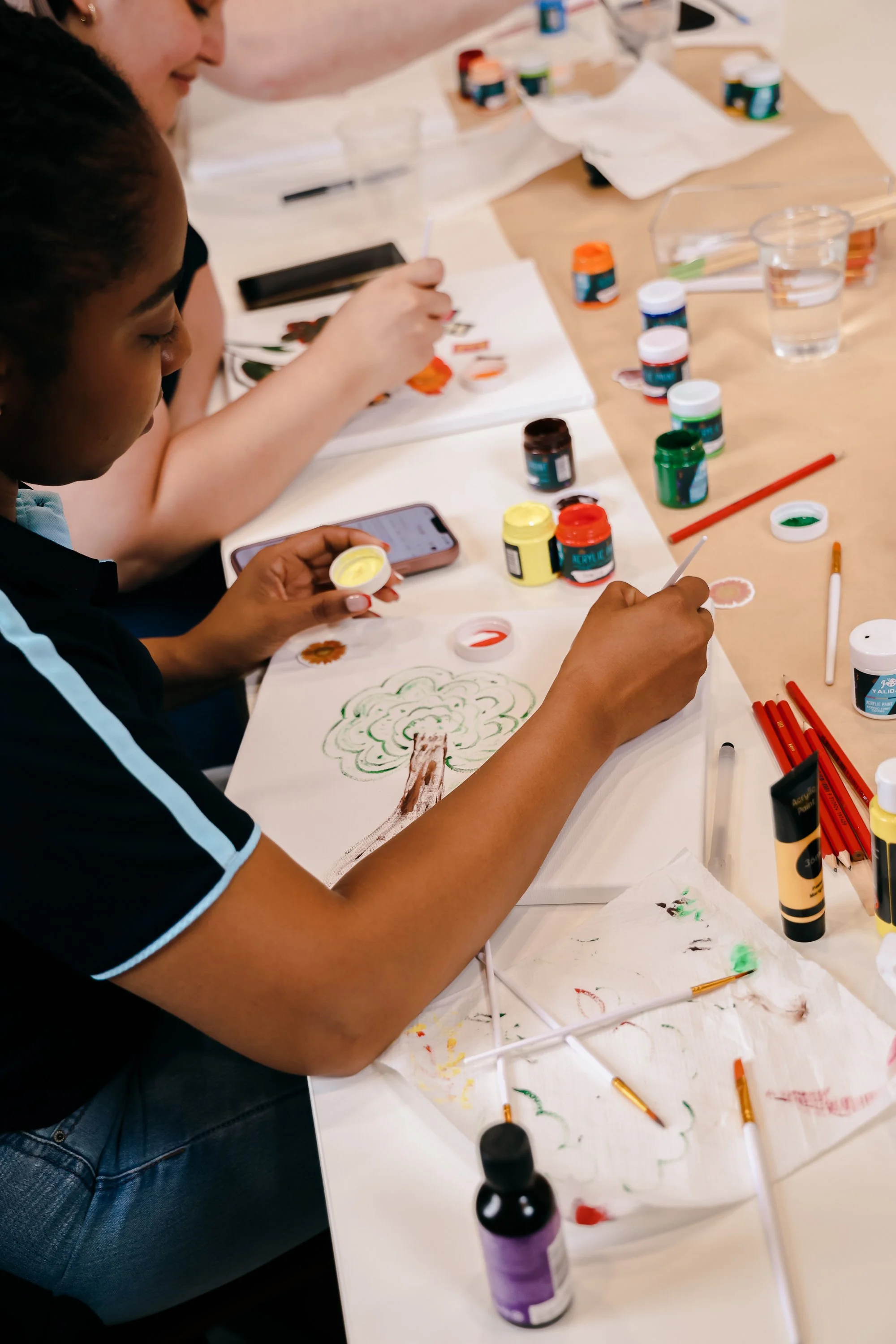 People painting and drawing at a table with various art supplies including paints, brushes, and paper, creating colorful artwork.