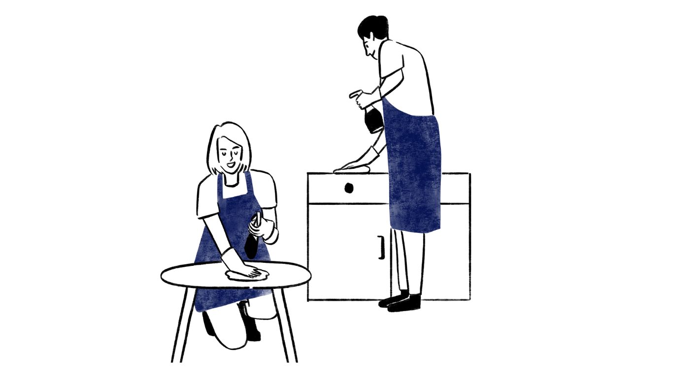 A woman sitting at a table washing dishes while a man stands on a cabinet cleaning the top of it.